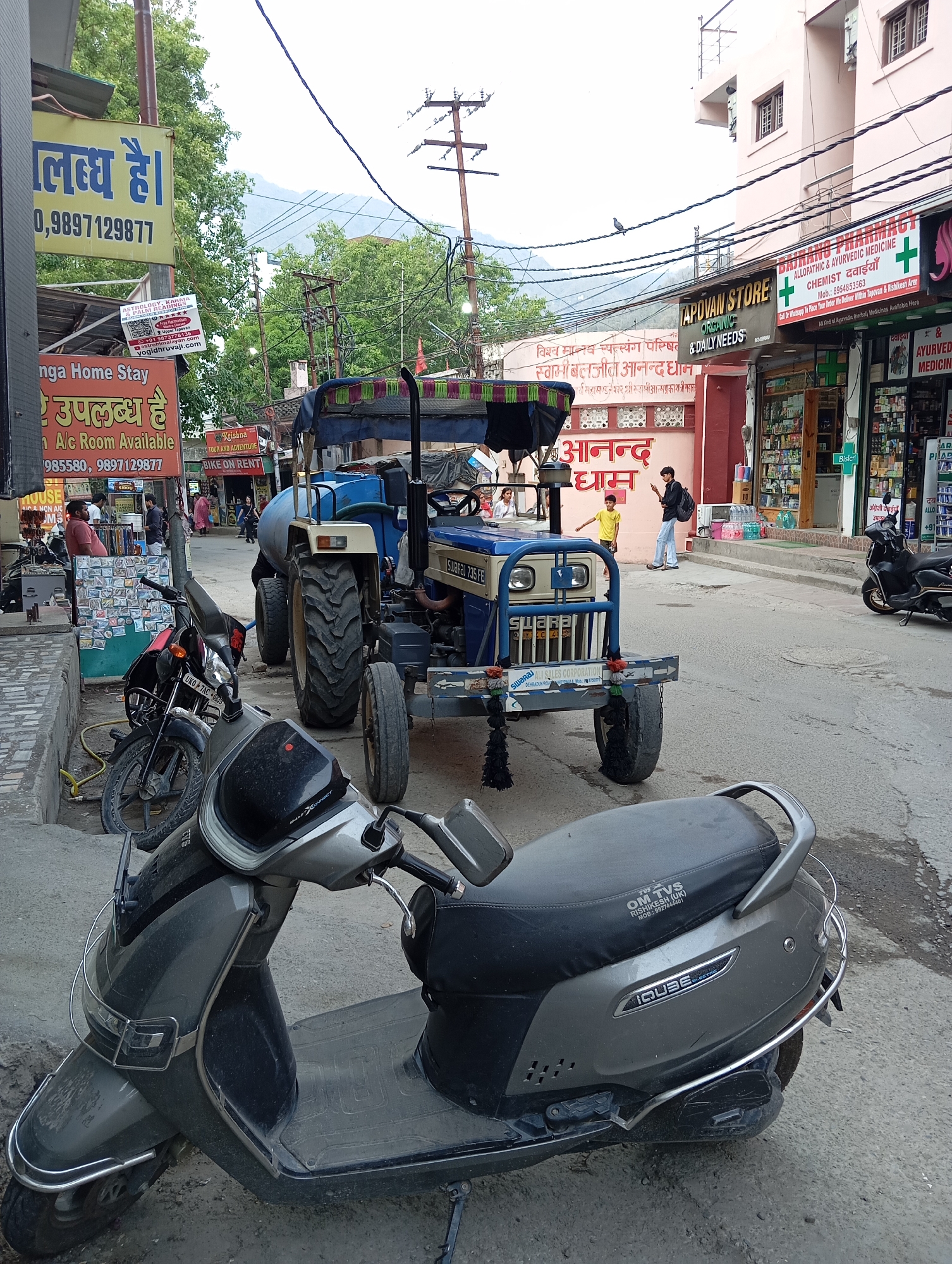 The image depicts a street scene in Rishikesh, India, dominated by a gray scooter parked in the foreground. Behind it is a blue tractor with "SWARAJ 735 FE" visible on its body. Several other motorcycles are parked nearby, partially obscured. In the background, people are walking on the sidewalk, some entering and exiting stores such as "APOVAN STORE" and a pharmacy. Signs for "Ganga Home Stay" and "Krishna Tour and Adventure" are also visible. The street is paved and moderately busy. Overhead, power lines crisscross the sky. The architectural style of the buildings is varied, with some appearing to be modern constructions and others more traditional.