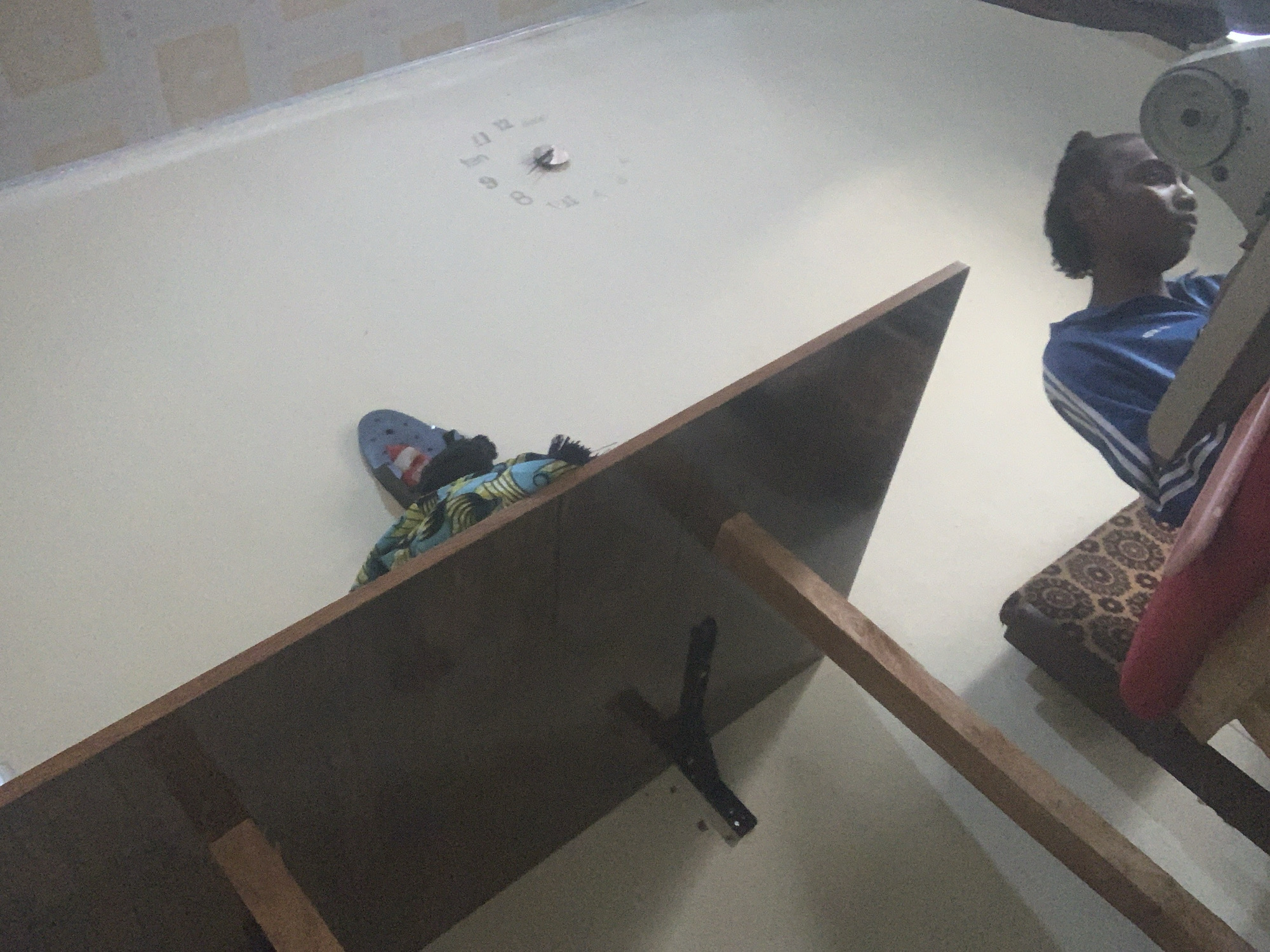 In Jauro Yinu, Nigeria, a person wearing a blue and white striped shirt is hunched over a sewing machine, seemingly focused on their work. The scene appears to be indoors, with a shelf or table overhead that has a patterned fabric and a shoe resting on it. A wall clock is visible on the ceiling, indicating the time. The overall lighting is dim, with the primary light source seemingly coming from the side, highlighting the person and the sewing machine. The background shows parts of furniture, including a patterned cushion.
