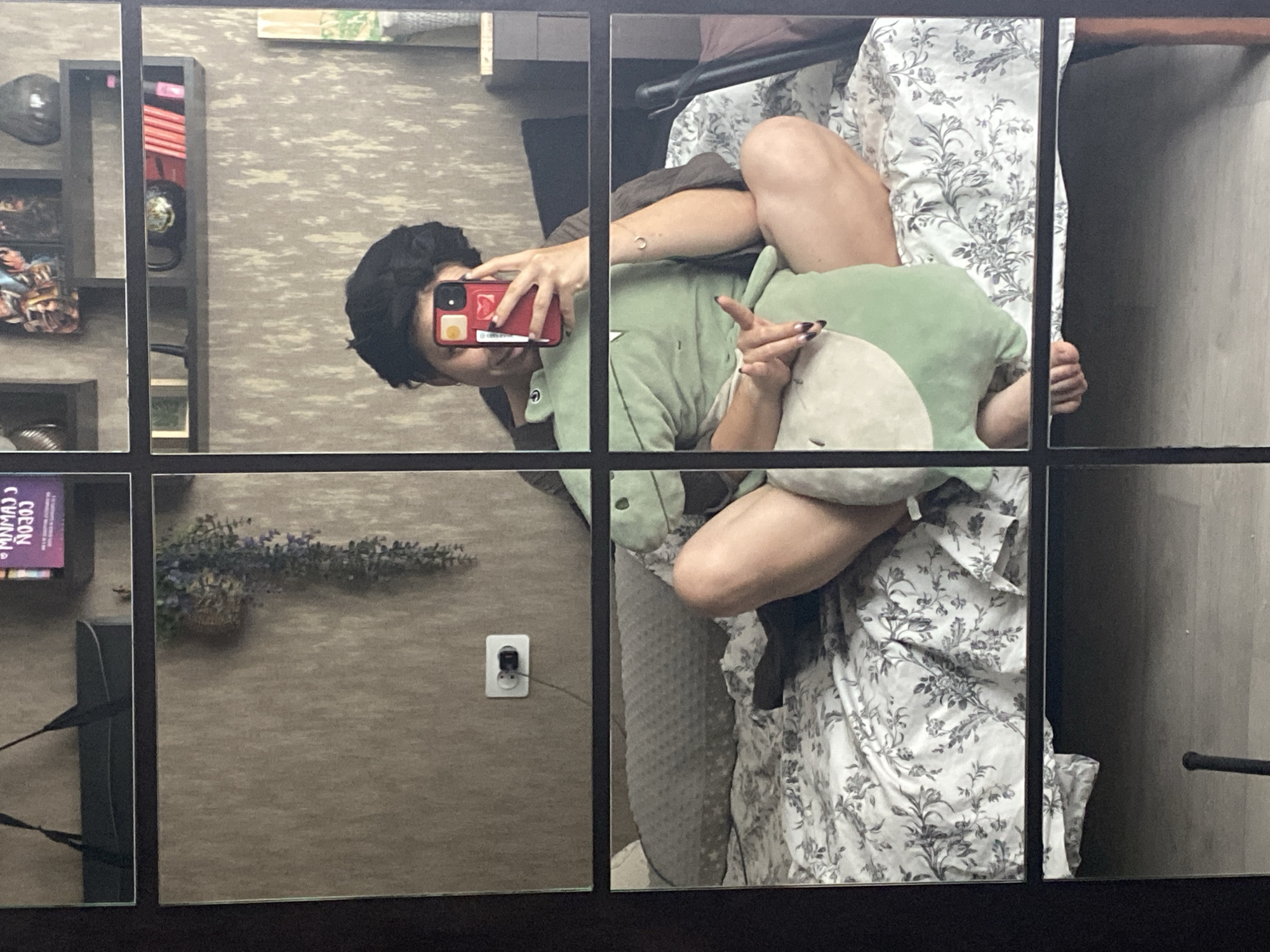 The image is a reflection in a mirrored wardrobe, depicting a person sitting on a bed. The person has short, dark, curly hair and is holding a red phone in their right hand, taking a selfie. They are wearing a dark colored shirt and are holding a large, light green stuffed dinosaur. The bed has a white duvet with a grey floral pattern. The background consists of a wall with a textured pattern, a hanging plant, and a white electrical outlet with a black plug. A shelf with books and decorative items is visible in the upper right section of the reflection. The overall setting appears to be a bedroom. The image does not contain any specific visual cues that would allow for the identification of the city of Chaikovsky, Russia, or any particular location within it.