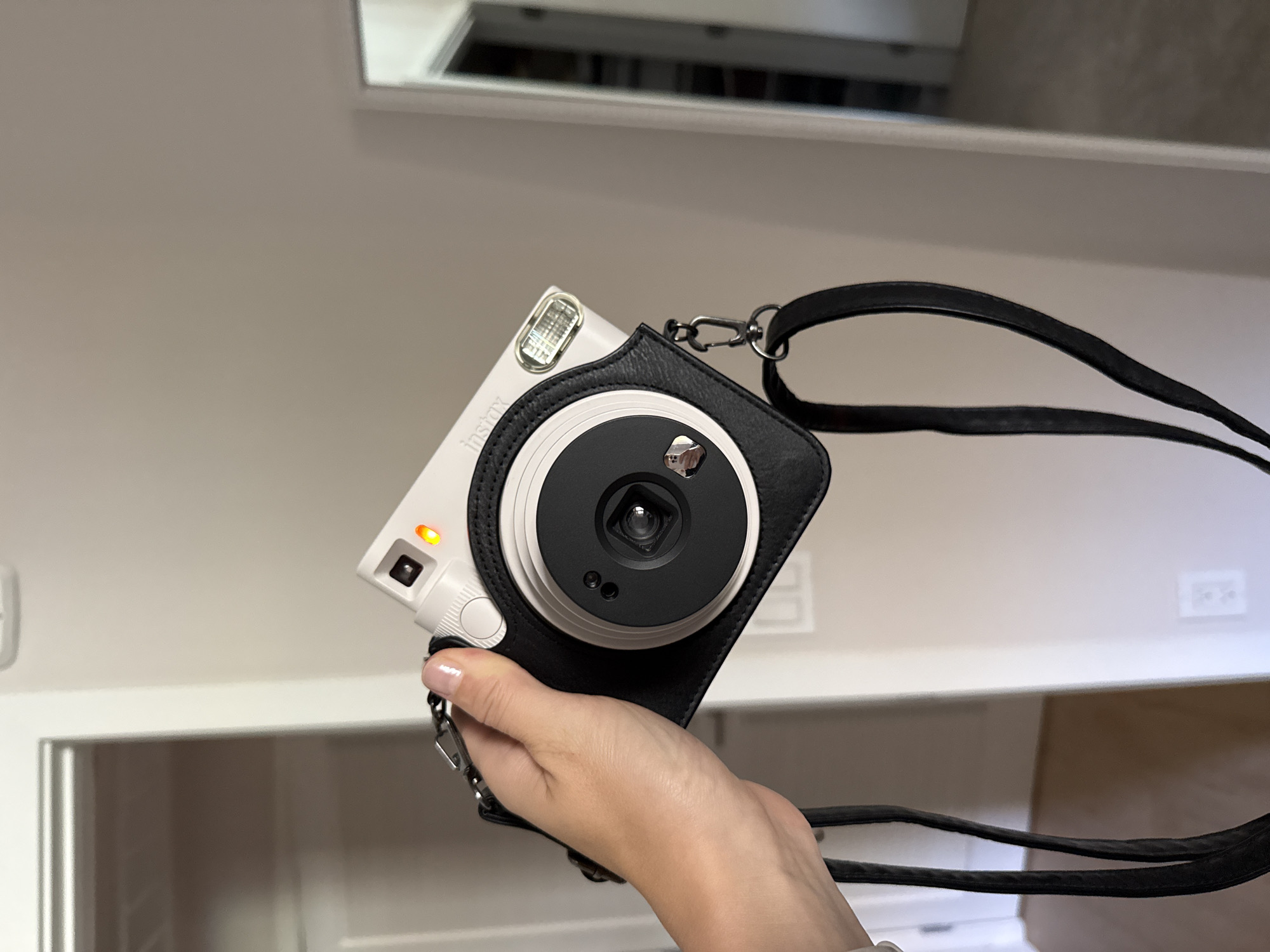 In Huntington Beach, California, a hand holds a white instant camera with a black leather case and strap. The camera is pointed downwards and to the left. The background consists of an interior room with neutral-colored walls, a doorway, and a wall outlet. The lighting is soft and diffused.