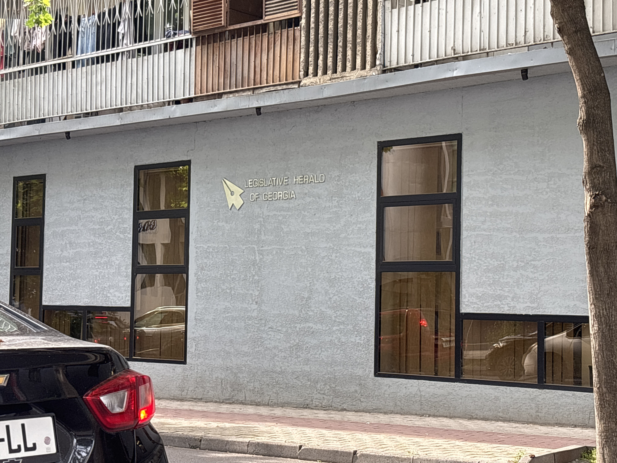 A building with a light grey stucco facade is shown in Tbilisi, Georgia. The building has several large, dark-framed windows, some of which reflect the surrounding environment and cars. Above the windows, there is a balcony with metal railings and laundry hanging. A tree trunk is visible on the right side of the frame. Text on the building reads "LEGISLATIVE HERALD OF GEORGIA" with a stylized yellow pen nib logo. A black car is parked in front of the building, and its license plate is partially visible. The street in front of the building is paved with bricks and asphalt.