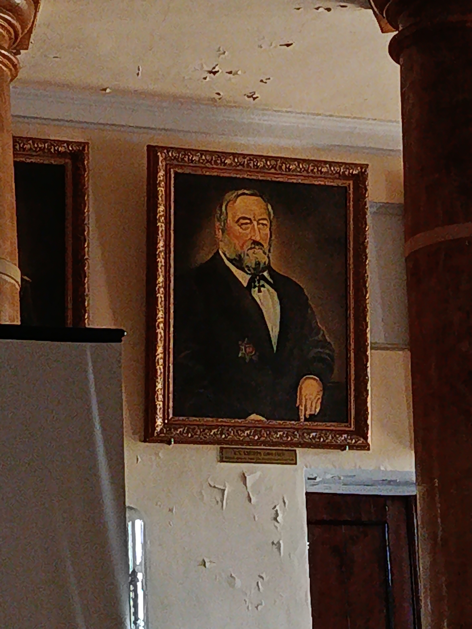 The image shows a portrait of a man with a beard and mustache, wearing a dark suit and a white shirt with a dark tie. He has a medal on his chest. The portrait is in a gilded frame and hangs on a wall. To the left of the portrait is another framed artwork, and to the right is a large pillar. The wall appears to be painted and shows signs of age and peeling paint. Below the portrait, a small plaque is visible. In the foreground on the left, a projection screen is partially visible, casting a shadow on the wall. The background shows a doorway and a window. The overall setting appears to be indoors, possibly a museum, gallery, or a historic building.