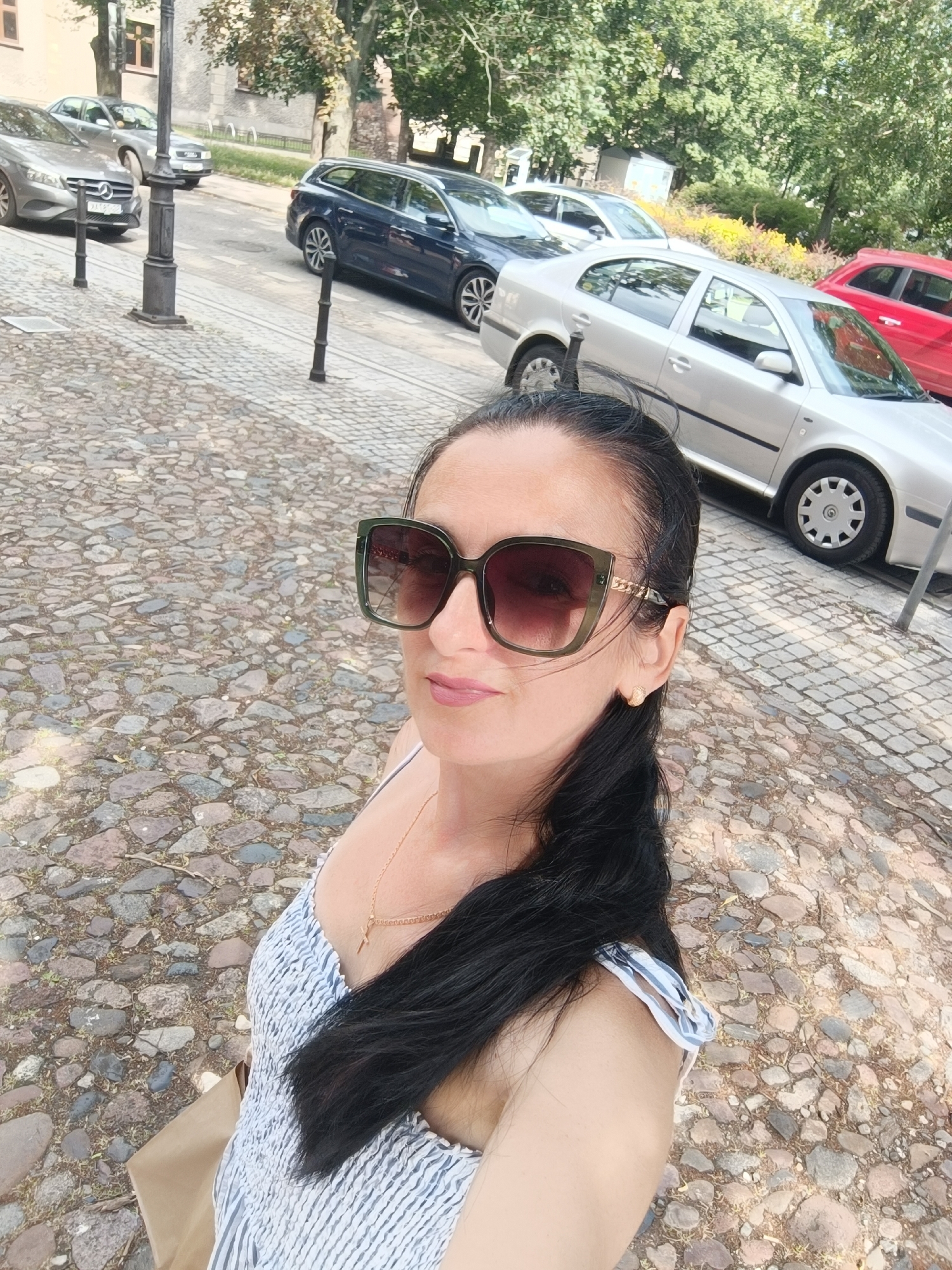 The image shows a woman with dark hair wearing large sunglasses and a striped dress, posing for a selfie outdoors. She is standing on a cobblestone street, with several cars parked along the street in the background. The environment appears to be a city street in Gliwice, Poland, likely in an older part of town given the cobblestones and architecture. Trees and buildings are visible in the background, suggesting an urban setting. The scene is captured on a sunny day.