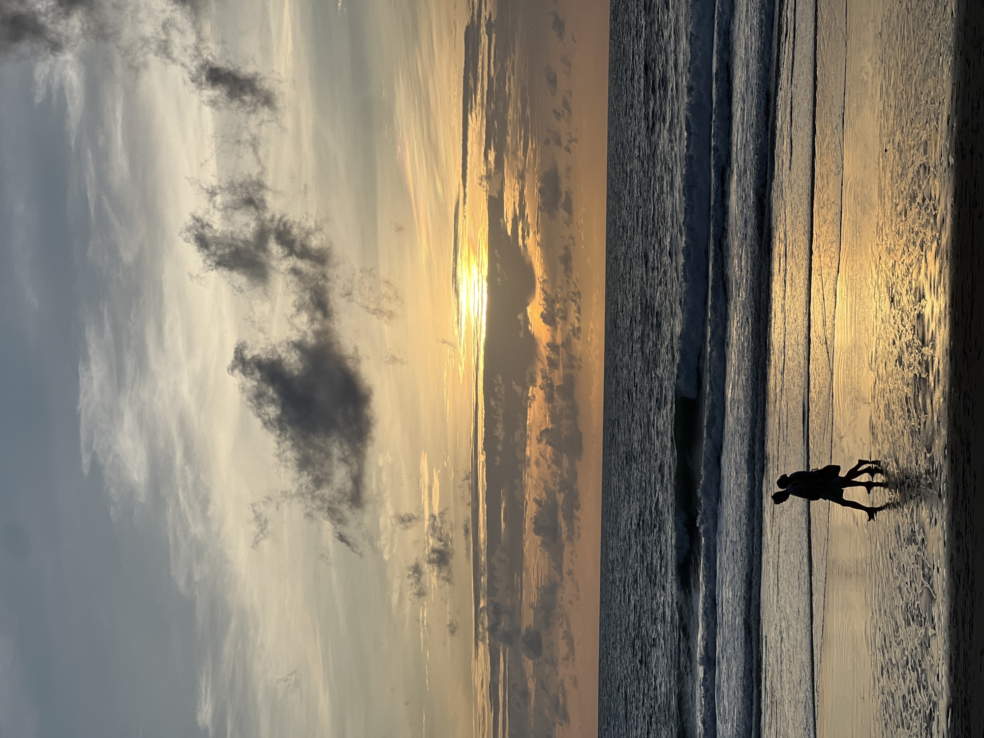 The image depicts a serene sunset scene at a beach in Choeng Thale, Thailand. Two silhouetted figures, a man and a woman, are walking hand-in-hand along the wet sand, their forms outlined against the shimmering golden light reflecting from the water. The ocean stretches out before them, with gentle waves rolling towards the shore. Above, the sky is a canvas of soft pastel colors, transitioning from a pale yellow and orange near the horizon where the sun is setting, to muted grays and blues higher up. Wispy clouds drift across the sky, some catching the last rays of sunlight and appearing as dark smudges, while others are illuminated with a soft glow. The overall atmosphere is one of peacefulness and romantic solitude, characteristic of a beach setting in Choeng Thale during twilight.
