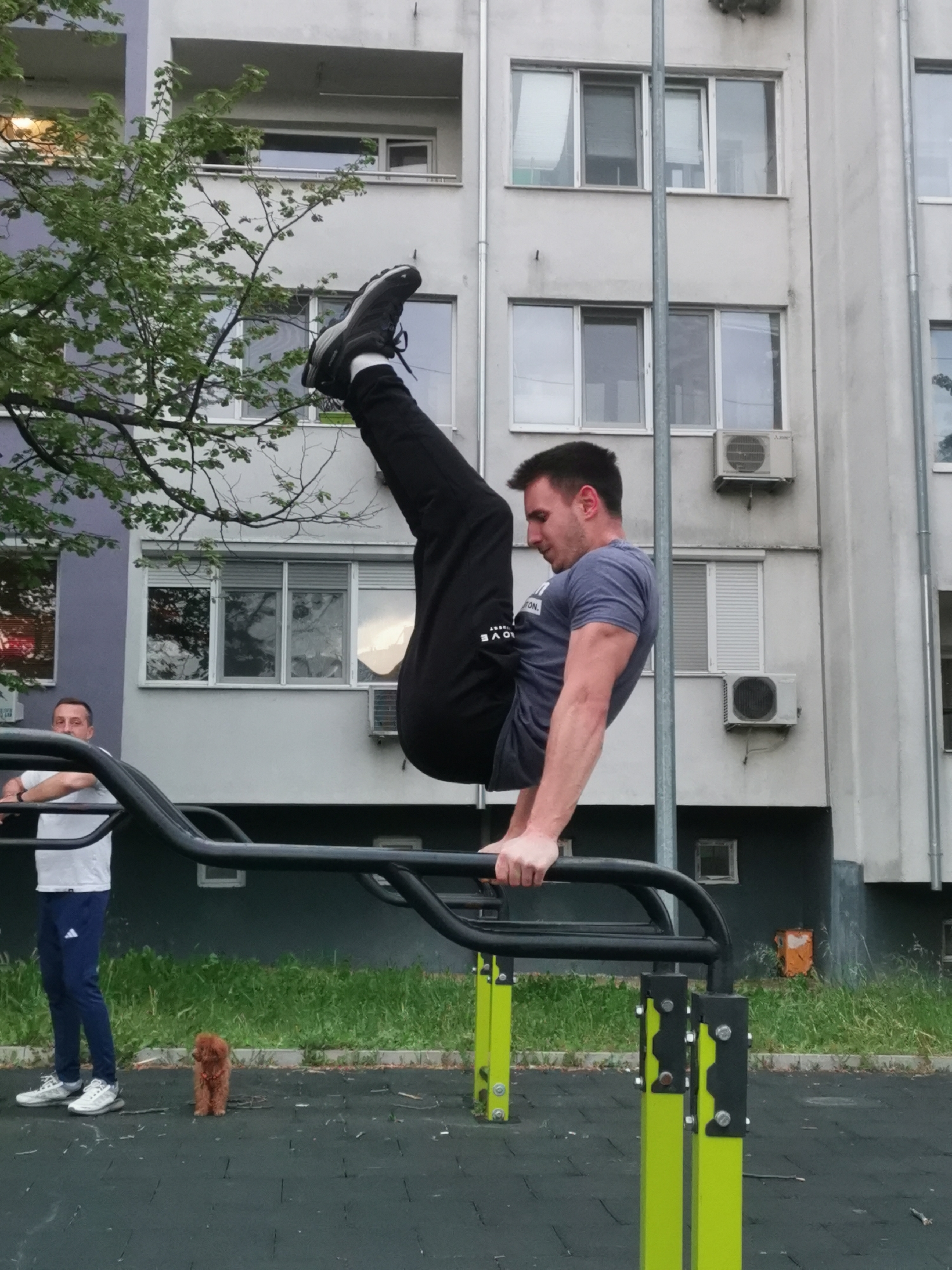 A man is performing a calisthenics exercise on a black metal outdoor gym apparatus with yellow accents. He's suspended above the ground, holding himself up with his arms straight and his body in a vertical position, legs raised high in the air. He is wearing a blue-gray t-shirt and black pants. Another man in a white shirt and blue athletic pants is standing nearby, seemingly observing, with a small brown dog standing in between the men. The setting appears to be an outdoor park or recreation area, with a multi-story residential building in the background. The building's exterior is a mix of gray and lilac with white window frames and several air conditioning units attached to the walls. The scene is located in the city of Burgas, Bulgaria.