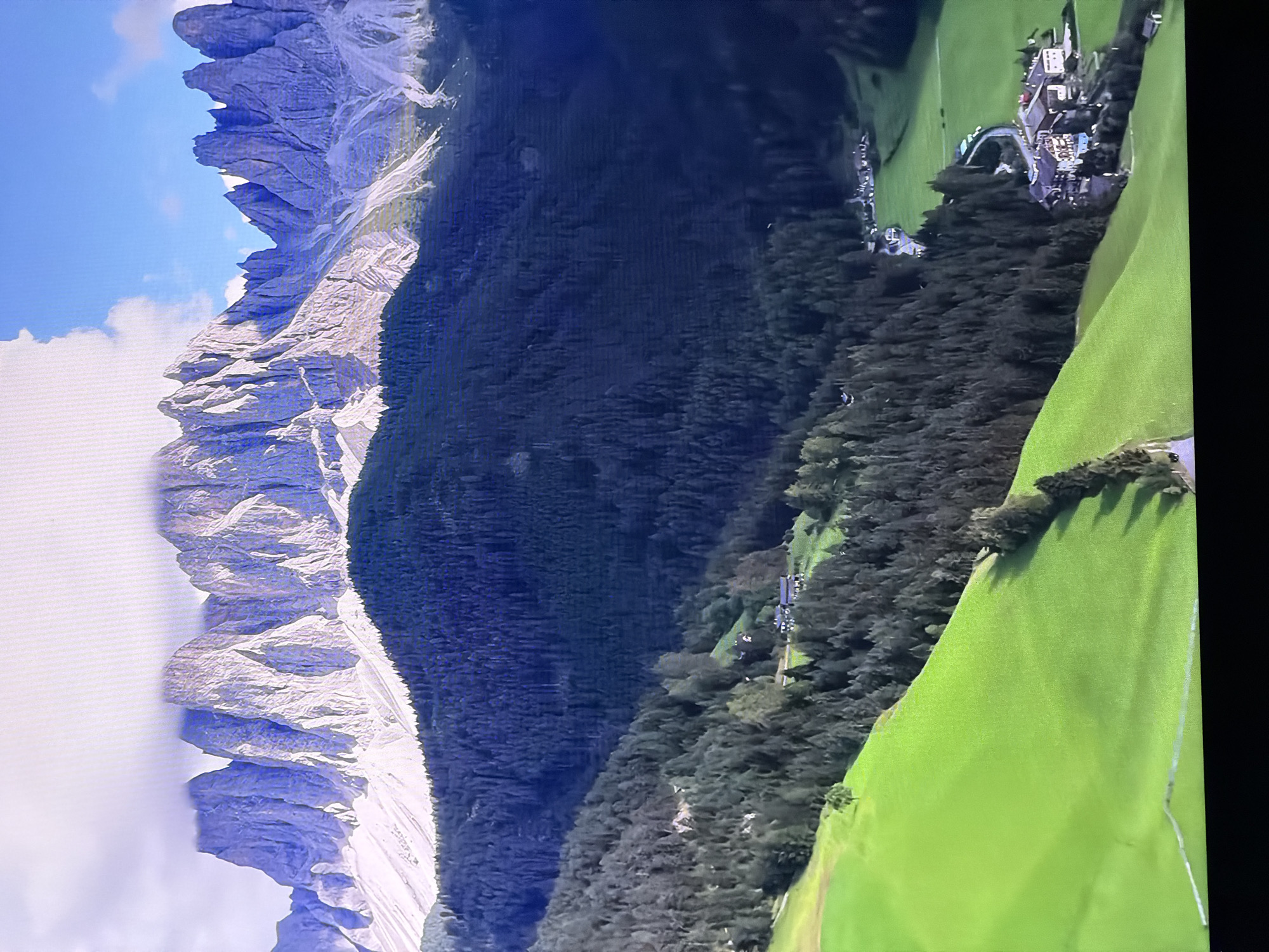 The image depicts a sweeping, panoramic view of a mountainous landscape. The foreground is dominated by a vibrant, undulating green meadow, dotted with a few scattered trees. A winding path is visible cutting through the meadow, with what appears to be a stone wall or fence running alongside it.
Further into the mid-ground, the landscape transitions to a dense forest of dark green conifer trees, carpeting the slopes of the mountains. Within the forest, a cluster of buildings, likely a small village or hamlet, is nestled amongst the trees. These structures feature red-tiled roofs and appear to be traditional alpine-style architecture. A road or track snakes through this settlement.
The background is defined by a towering mountain range, characterized by jagged, rocky peaks. These mountains are illuminated by sunlight, casting shadows that emphasize their dramatic forms. A portion of the mountain peaks is shrouded in a soft, white cloud formation. The sky above is a clear, bright blue with a few wispy clouds.
The image is a static, landscape photograph. There are no people or animals visible. The scene conveys a sense of natural beauty and tranquility.
The location is identified as the city of Chirchik, Uzbekistan.