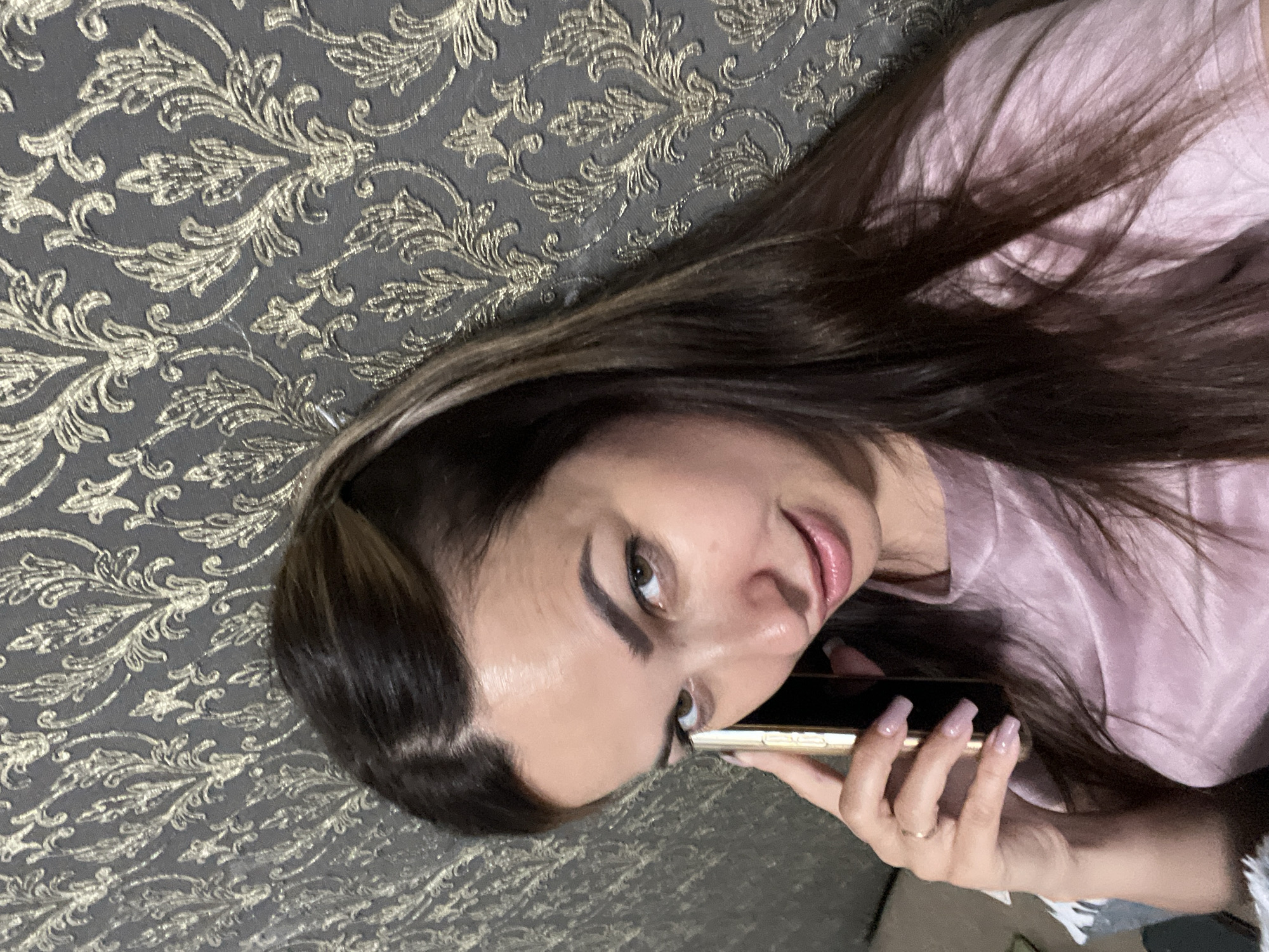 A young woman with long, dark brown hair is pictured in a close-up shot, holding a smartphone to her right ear with her right hand. She is wearing a pink, silky shirt. The background features a textured wallpaper with an ornate, golden damask pattern on a dark gray base. She is looking towards the camera with a slight smile. Her left hand is resting on the phone. The lighting appears to be soft and indoor. The scene does not provide any information about its location in the city of Unknown, Unknown.