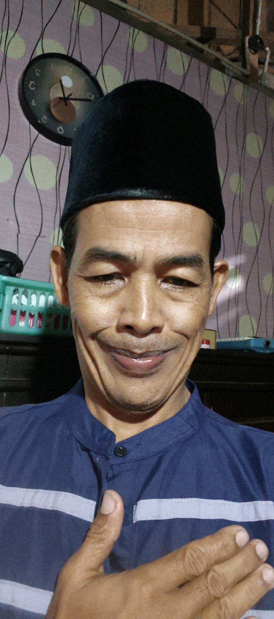A man in a black peci and a blue shirt with horizontal stripes is looking at the camera with a wide smile. His right hand is raised to his chest, with his thumb extended upward in a "thumbs-up" gesture. Behind him, a wall with a patterned wallpaper is visible, along with a round clock on the wall. To the left of the man, a green plastic container with compartments can be seen. The overall setting appears to be an indoor environment, possibly a home in Muaro Jambi, Indonesia.