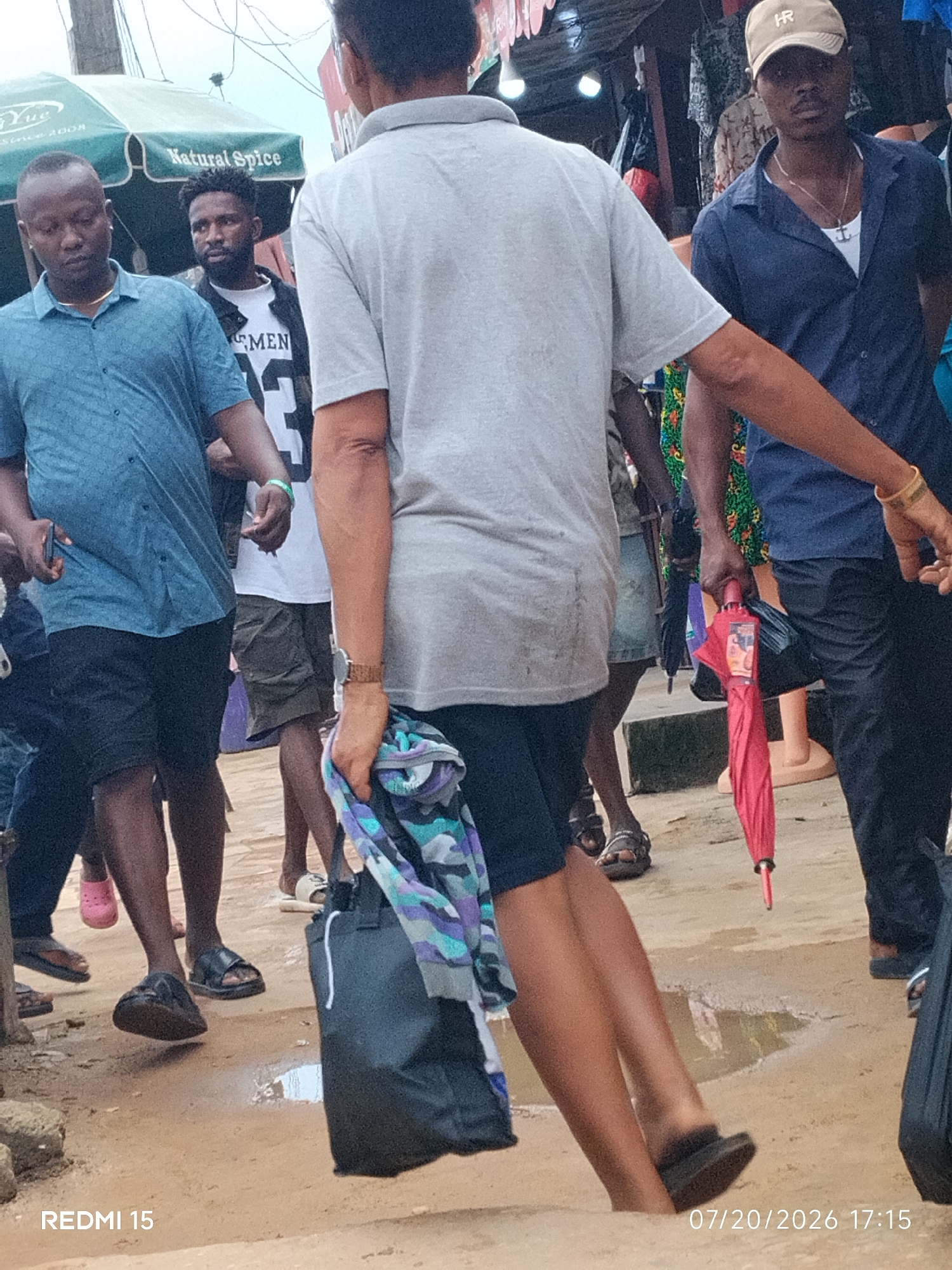 The image depicts a street scene in Umuode, Nigeria. In the foreground, a person walks away from the camera, carrying a black bag and a folded scarf. Another person is visible to their right, wearing a dark blue shirt and a cap. To the left, a man in a blue shirt holds a phone. In the background, several other individuals are present, along with market stalls and a green awning. The ground appears to be a dirt path with some puddles. The overall atmosphere suggests a busy marketplace.