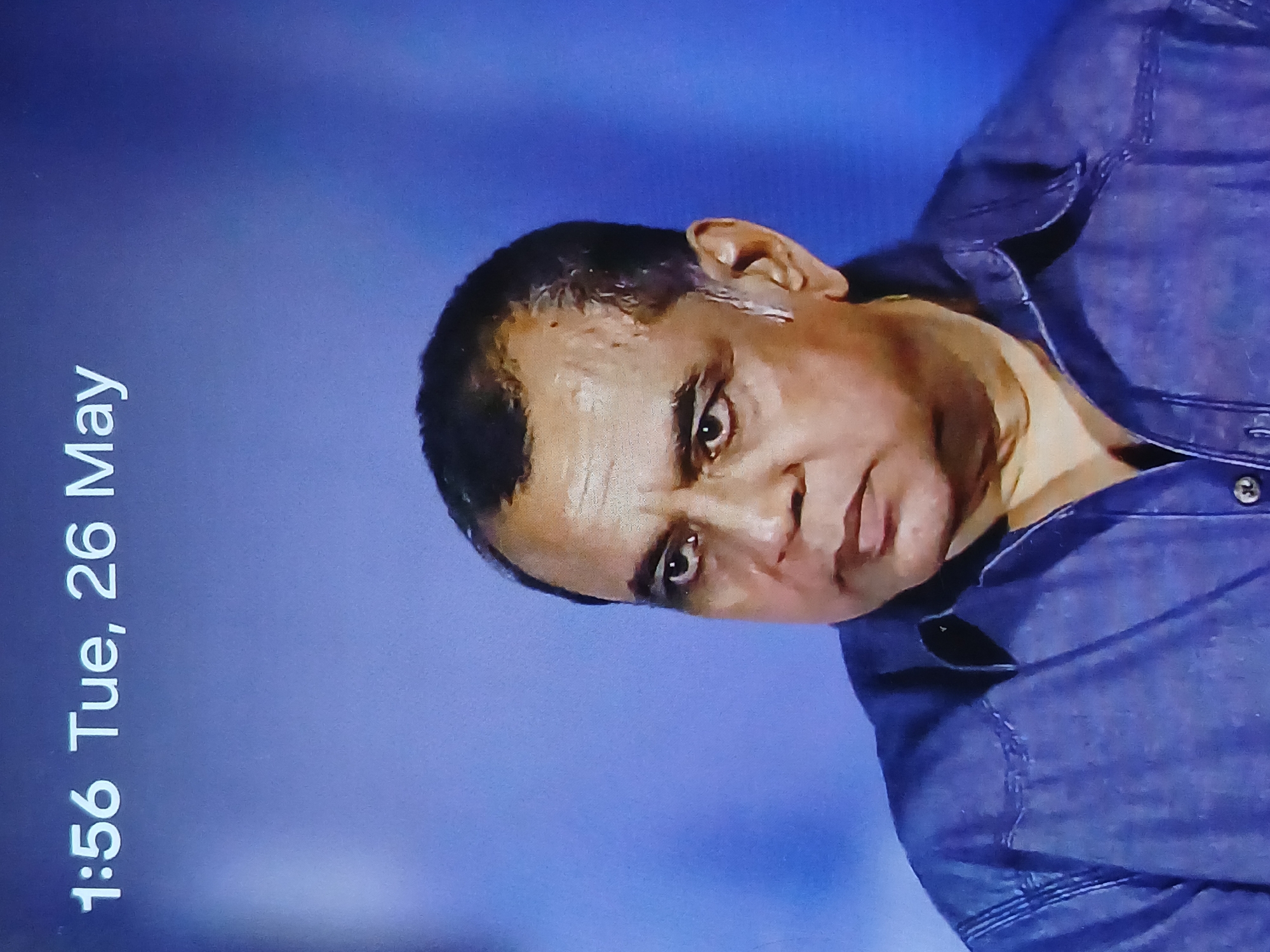 The image depicts a man wearing a dark blue button-down shirt, set against a light blue background. The date "Tue, 26 May" and the time "1:56" are visible on the left side of the image. There is no information available to determine the location in Bengaluru, India.