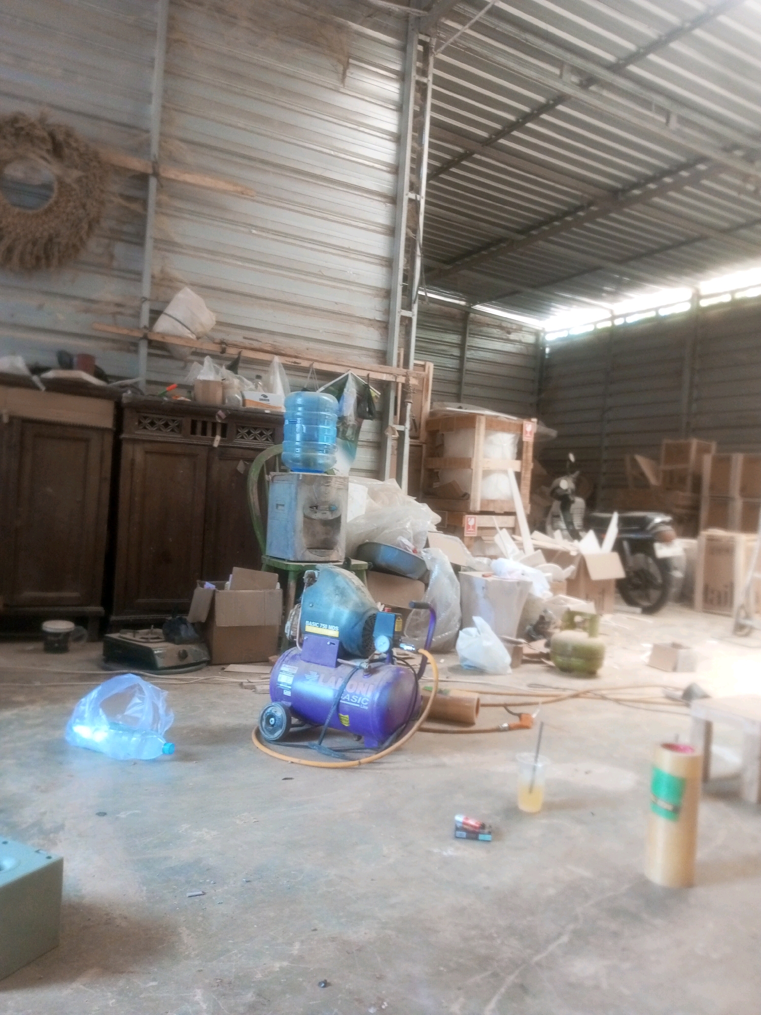 The image depicts a cluttered storage area within a workshop or warehouse located in Pajangan, Indonesia. The environment appears to be a metal-clad building with a corrugated metal roof, typical of industrial or semi-industrial structures in the region.
In the foreground, a purple air compressor with yellow hosing is prominent. To its left, a plastic bag containing a water bottle lies on the concrete floor. Further left, a small green stovetop and a dark object are visible. Behind the air compressor, a green propane tank is situated.
To the left of the compressor, a wooden cabinet or dresser is positioned, with another similar unit to its left. Above these cabinets, a blue water cooler dispenser with a large water jug is placed on a light blue stand. Behind the water cooler, there are stacks of boxes and various other items, including plastic bags and what appears to be a large blue tub.
In the background, a black motorcycle is parked. Various wooden crates and cardboard boxes are stacked throughout the space, suggesting ongoing operations or storage of goods. On the far left wall, a circular woven decoration hangs. The overall impression is one of a functional, albeit disorganized, workspace filled with equipment, supplies, and a motorcycle. The lighting suggests it might be daytime, with some light filtering in from the sides of the building.