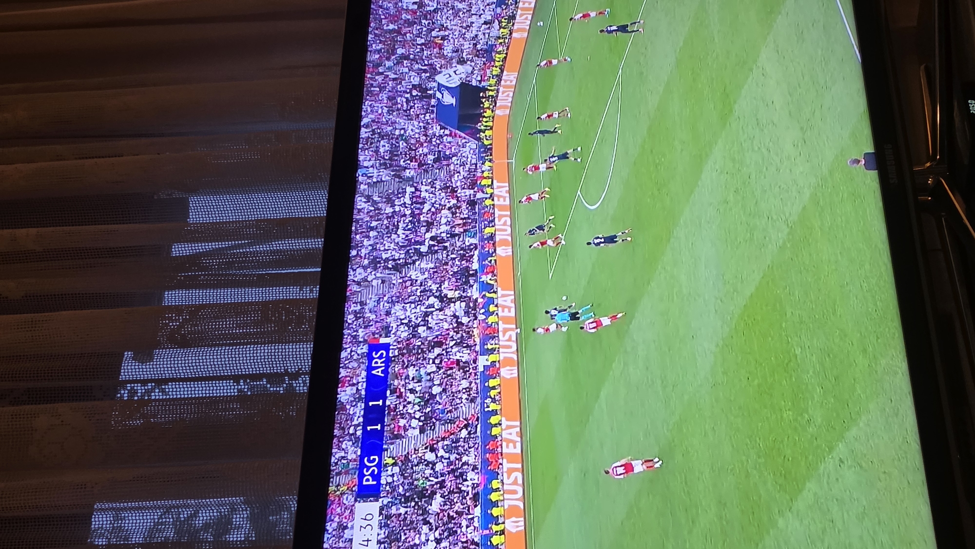 The image shows a television screen displaying a soccer match. The score is PSG 1, ARS 1. The image is rotated counter-clockwise. The match appears to be in progress, with players visible on a green field. Spectators fill the stadium in the background. "JUST EAT" is displayed on the side of the field. The TV is located indoors, in front of window blinds. There is no evidence of a location in Rogachevo, Bulgaria.