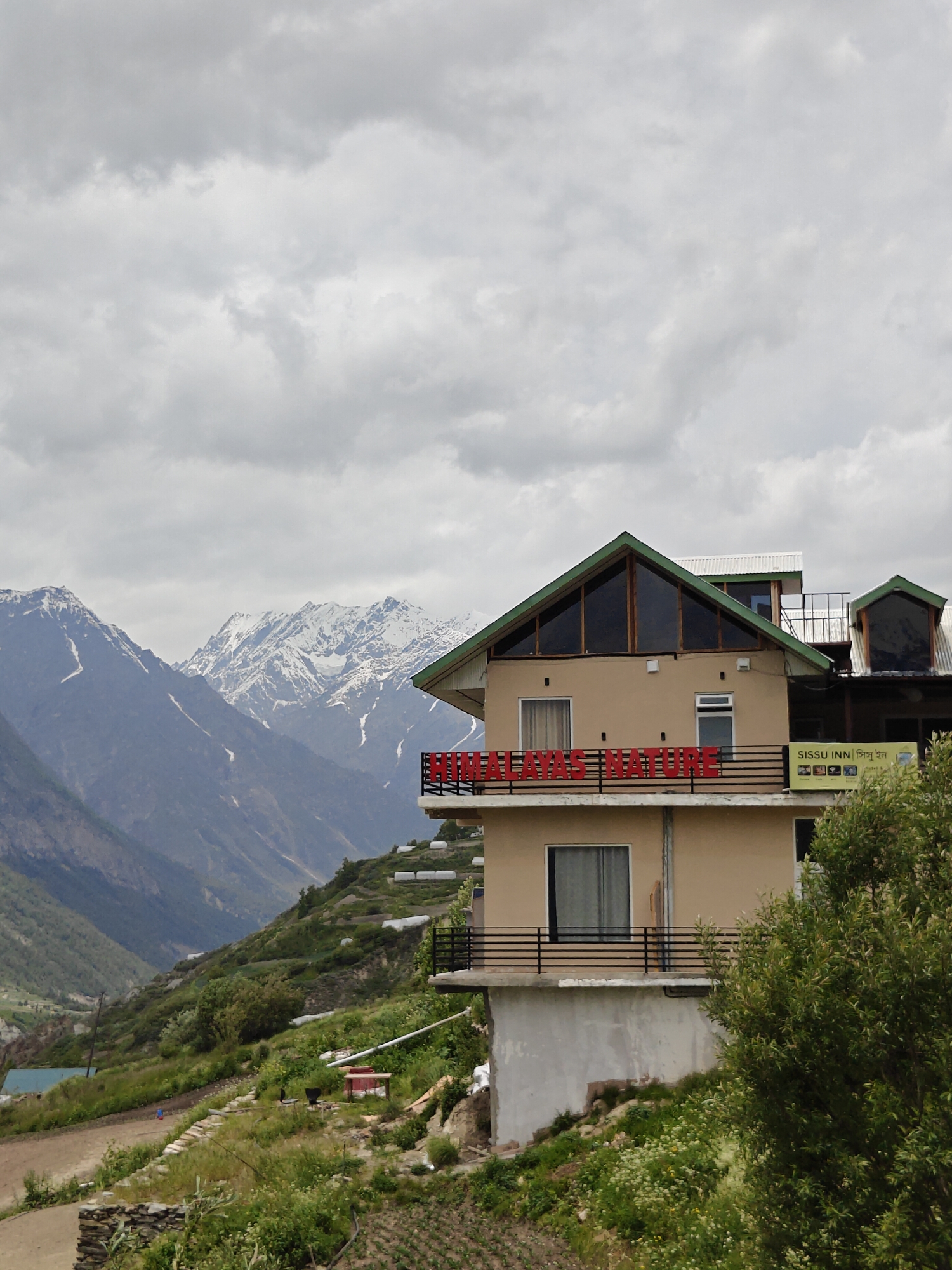 A hotel named "Himalayas Nature" is visible in the foreground. The hotel is a multi-story building with a beige exterior and green triangular roofs. Large windows with dark frames are present on the top floor. A balcony with a red railing runs along the middle floor. To the right of the hotel, a yellow sign with text in English and what appears to be an Indian script reads "SissU INN". Snow-capped mountains dominate the background. The sky is overcast with grey clouds. The hotel is situated on a hillside covered with vegetation.