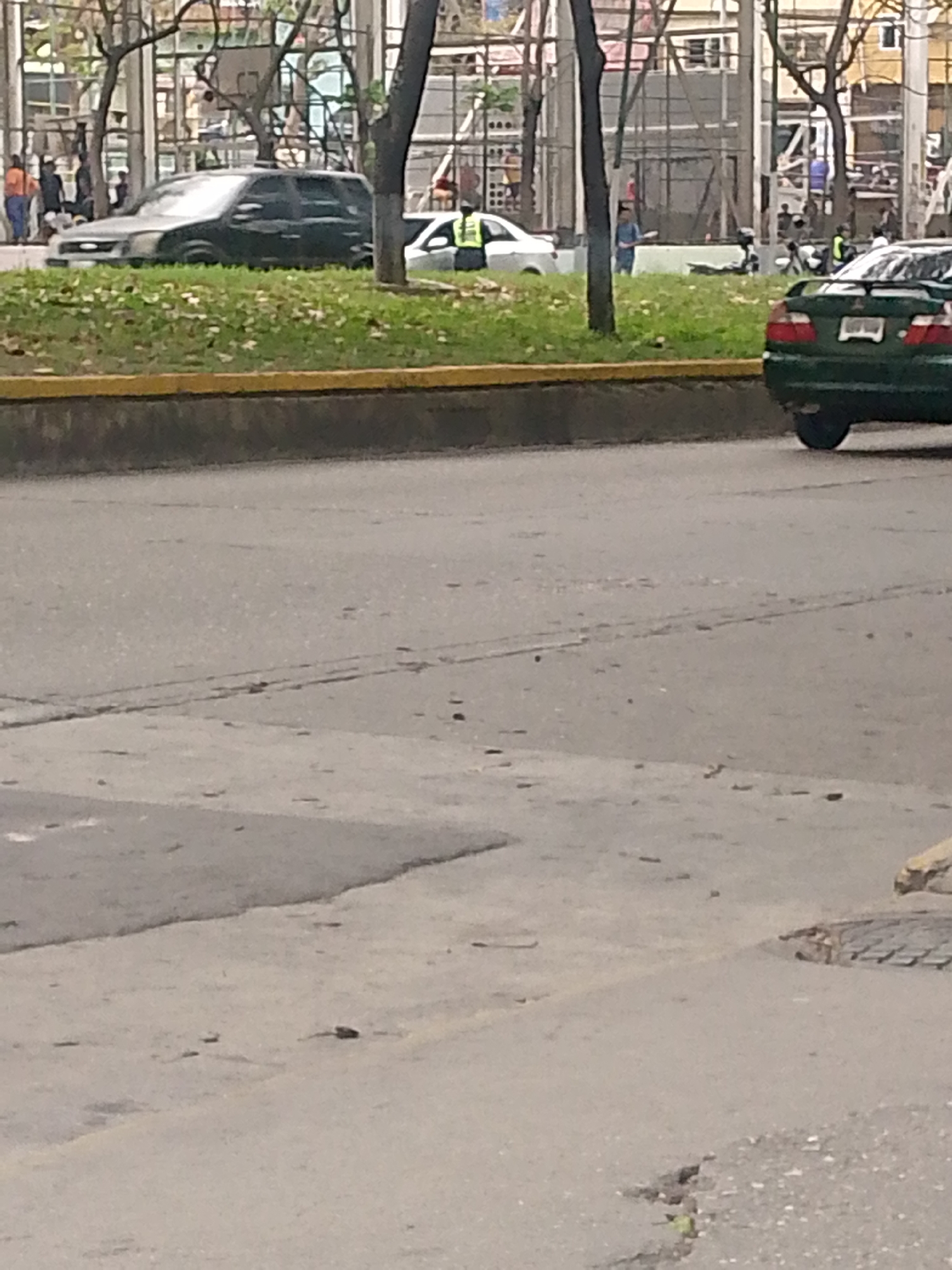 The image shows a street scene in Caracas, Venezuela. In the foreground, there is a paved street with cracks and a manhole cover. In the background, there is a raised green strip with trees. Behind the trees, there is a sidewalk with people, including some wearing orange vests. There are several vehicles visible, including a dark SUV, a white sedan, and a green sedan. A person wearing a vest with yellow stripes is visible next to the white sedan. A construction site or a building facade is visible further in the background.
