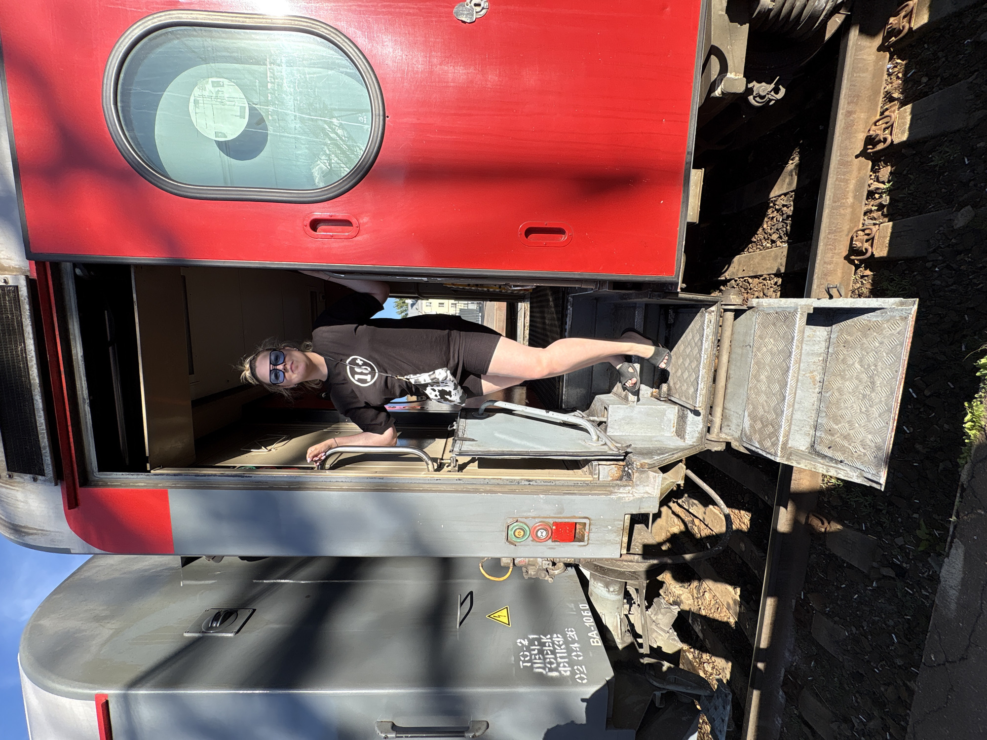 A woman stands in the passageway between two railcars. The passageway is between a red railcar above and a grey railcar below. The woman wears sunglasses, a black t-shirt, and black shorts. She is leaning with her left hand on a horizontal railing and her right foot is visible on the steps leading into the passage. Railroad tracks are visible between the railcars. The writing “TO-2” and “AE34-1” in Cyrillic is visible on the grey railcar. The scene is likely in Saratov, Russia, based on the Cyrillic text.
