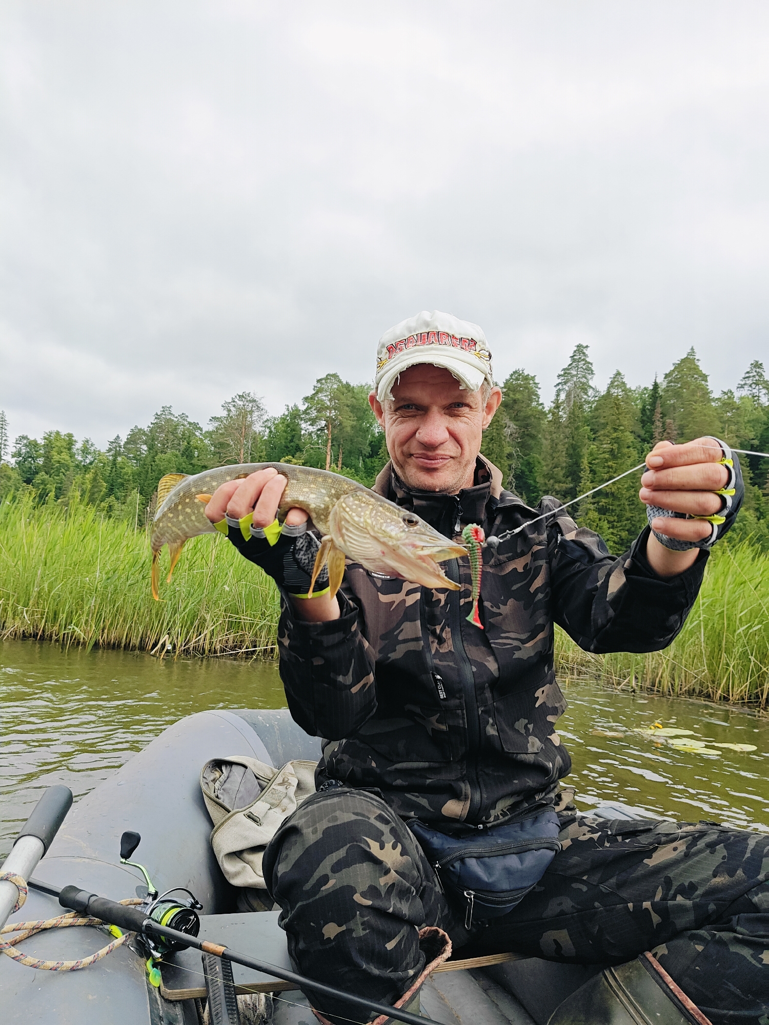 A man, wearing a camouflage jacket, camouflage pants, and a white baseball cap, is sitting in an inflatable boat. He is holding a fish in his left hand and a fishing line with a lure in his right hand. The boat is on a body of water, with tall green grass and trees in the background. The sky is overcast. The man appears to be in the city of Detskii Dom (Bashenino), Russia.