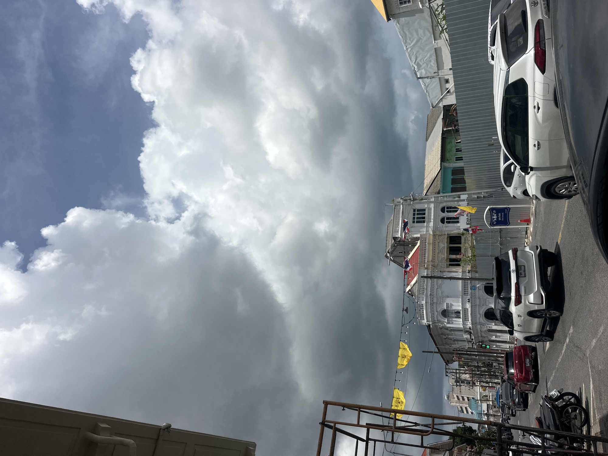 The image depicts a street scene in Phuket City Municipality, Thailand, on a day with a dramatic sky filled with a mix of dark storm clouds and bright white cumulus clouds. The foreground features a paved road with several vehicles, including a white SUV in the right lane, a red car in the left lane, and other cars further down the street. To the left, there are parked motorcycles.
The architecture of the buildings lining the street is a notable feature. On the left, a building with a white facade and ornate detailing is visible. Further down the street, more buildings with pitched roofs and colonial-style elements are present. Some of these buildings appear to be old, with distinctive window designs and balconies. Yellow banners are strung across the street between poles, and strings of lights are also visible. A metal scaffolding structure stands on the left side of the road.
A metal fence separates the street from a construction or renovation area on the right side of the image. The overall impression is of a city street in a tropical location, with the weather contributing a sense of anticipation or impending change. The lighting suggests it is daytime, with diffused sunlight filtering through the clouds.