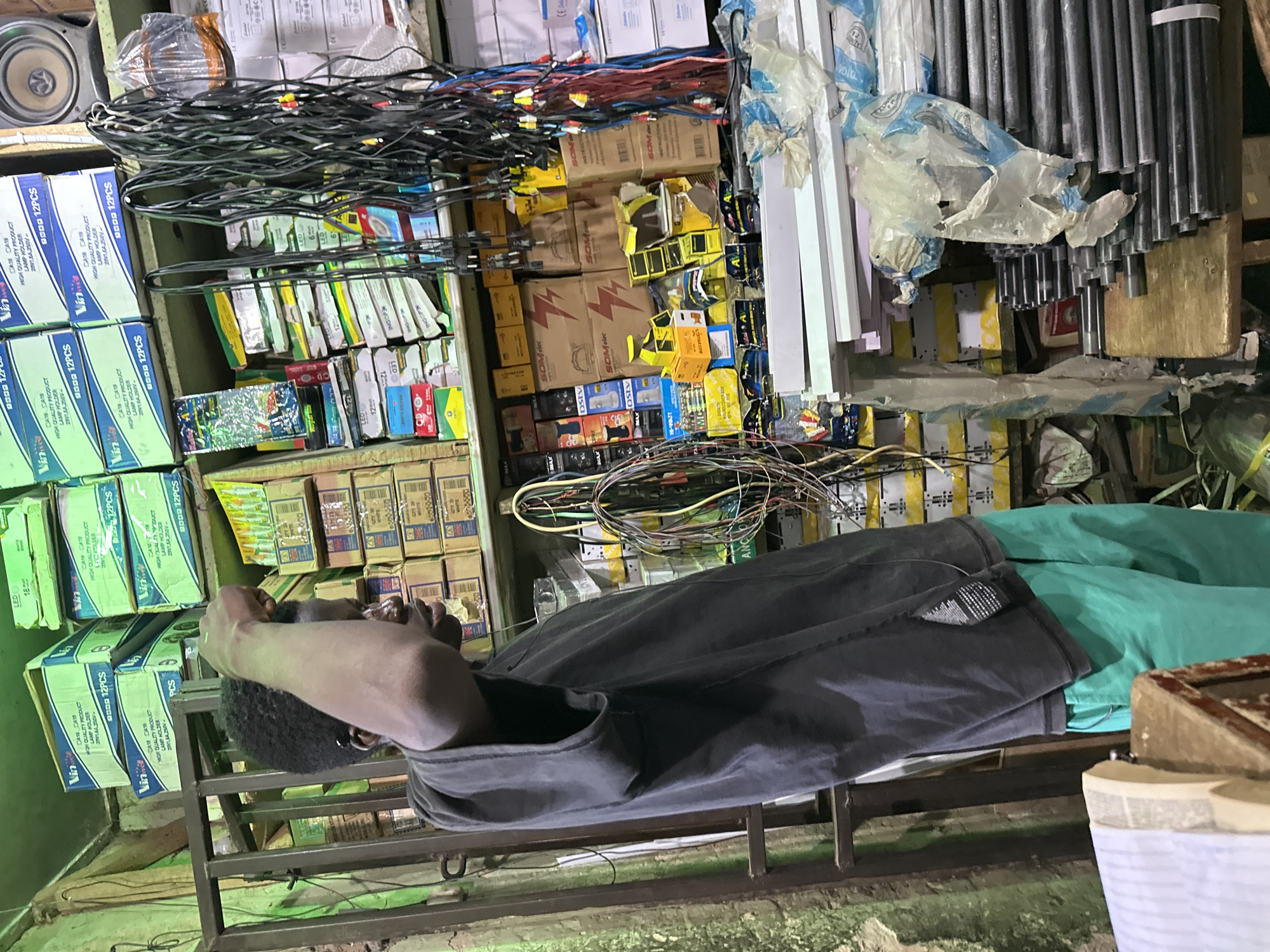 The image depicts a young Black male, likely a teenager, standing in a cluttered shop in Narayi, Nigeria. He is wearing a dark gray t-shirt and bright green shorts. His right arm is raised to his head, and he appears to be looking upwards and to the right. The background is filled with shelves stacked with various electronic components and supplies, including boxes of lamp holders, cables, and what appear to be lighting fixtures. Some items are neatly organized, while others are in disarray, with cables hanging loosely. The shop's walls are textured and appear to be made of concrete, with some signs of wear and tear. The lighting is somewhat dim, suggesting an indoor environment. In the foreground, to the left, a portion of a book or ledger is visible on a wooden surface.
