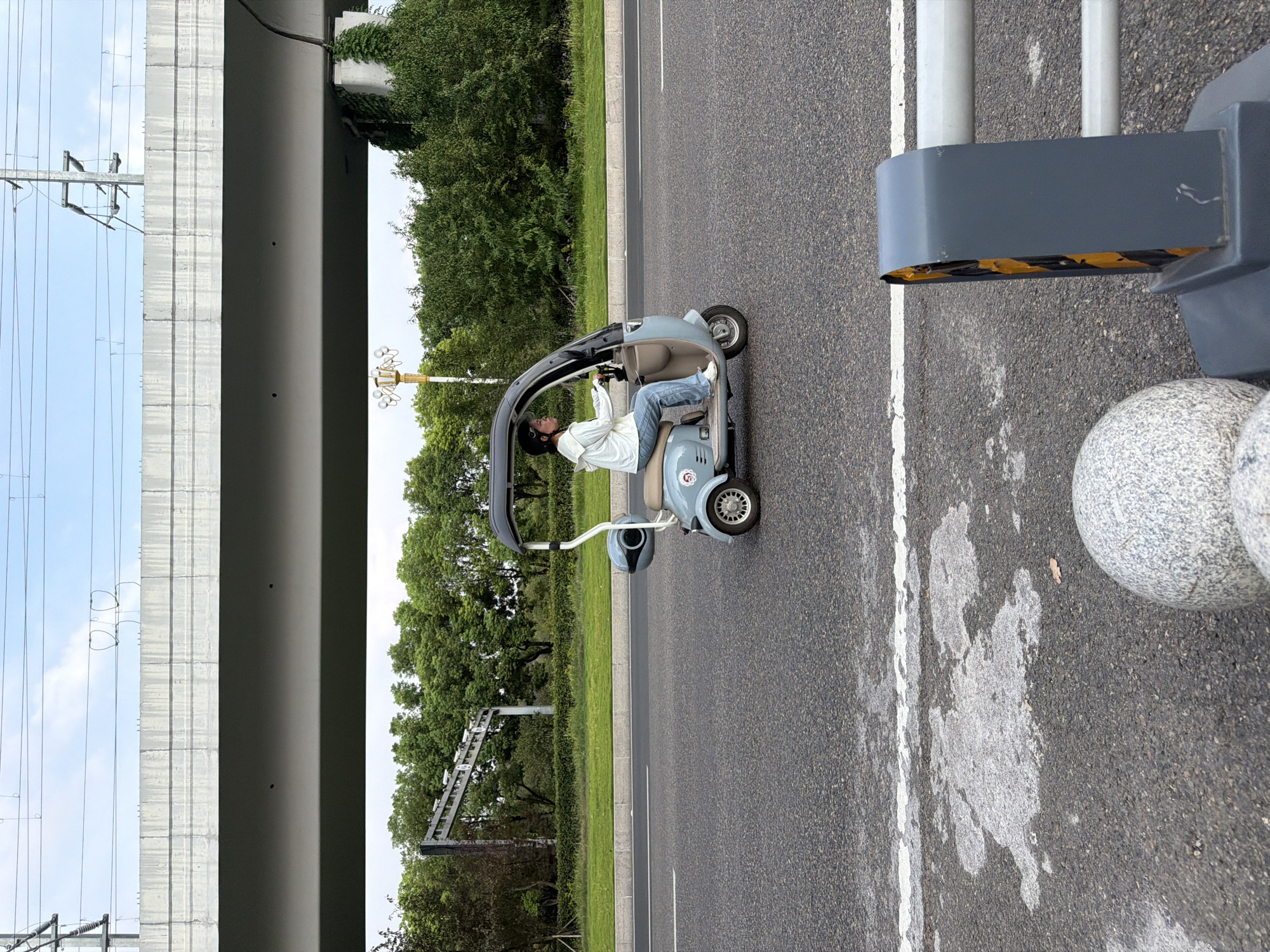 In the city of Jiangjin, China, a woman drives a light blue, three-wheeled scooter with a canopy on a paved road. She is wearing a white hooded jacket and blue jeans. The road has a white line marking its edge. In the background, a concrete overpass runs horizontally across the image, with overhead power lines and poles. Beyond the overpass, there are trees and greenery. In the foreground, partially visible, are parts of a gray barrier with yellow and black striped reflective tape, and two large gray spherical objects. The sky is cloudy.