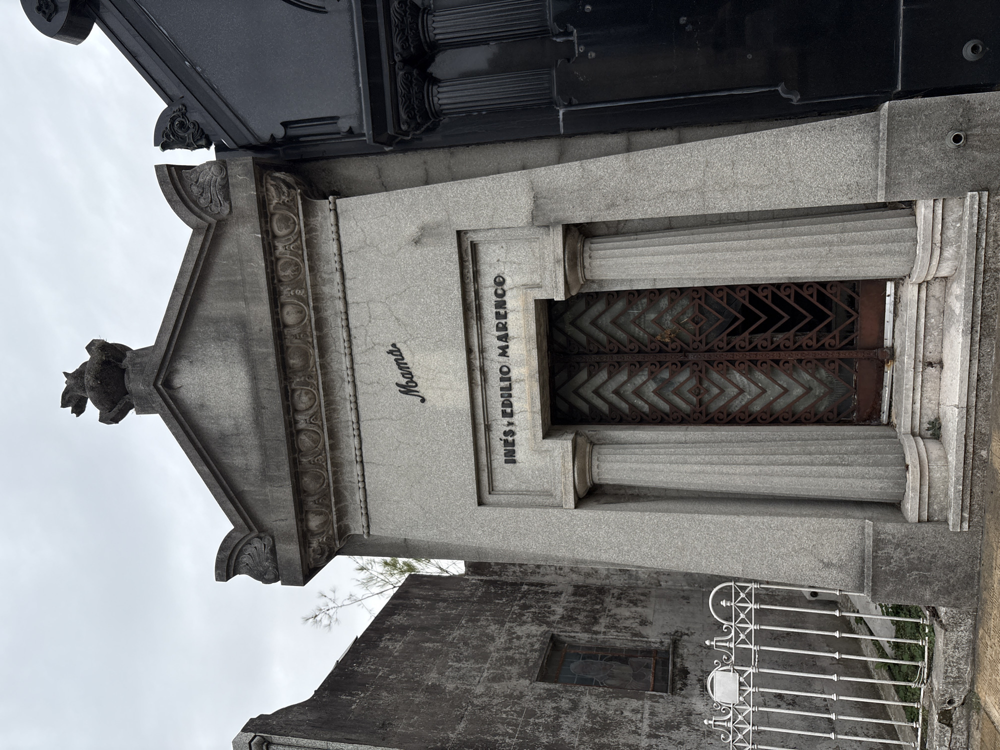 The image depicts a weathered grey granite mausoleum, identified as belonging to "INES Y EDILIO MARENCO" and inscribed with "Mamá" in cursive script above, located within the Recoleta Cemetery in Buenos Aires, Argentina. The mausoleum features a central double-leaf metal door with a geometric diamond pattern, flanked by two fluted stone columns, and accessed by two stone steps. The door is dark reddish-brown and appears slightly ajar. Above the main inscription, a decorative cornice supports a pediment-like structure topped by an indistinct, dark sculpture, possibly an animal.
To the immediate left of the central mausoleum stands a tall, rough-textured grey concrete or stone wall, belonging to an adjacent structure, featuring a single small, rectangular window with darkened or stained glass. A white, ornate metal fence, showing signs of wear and slight damage, runs along the base of this wall, with sparse green vegetation visible behind it. To the right, a portion of another mausoleum, constructed from dark, possibly black, polished stone, is visible, featuring ornate dark columns. The sky is overcast. No people are present in the scene.