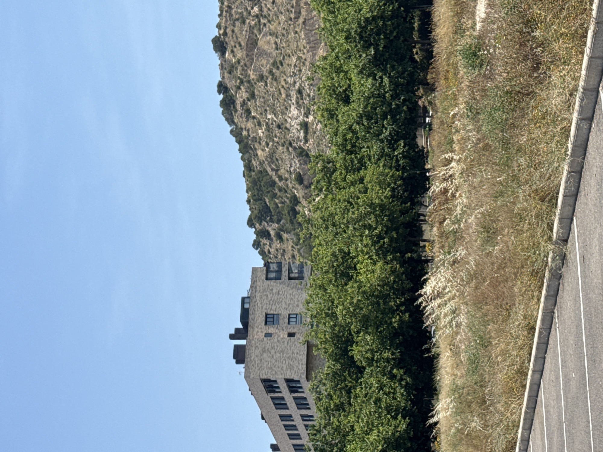 The image shows a building with a mountain in the background and a road to the side. The location is Unknown, Unknown. The building is tall and narrow, with many windows. The mountain is rocky and barren. The road is paved and has white lines. There are trees between the building and the mountain, and grass to the side of the road. The sky is blue.