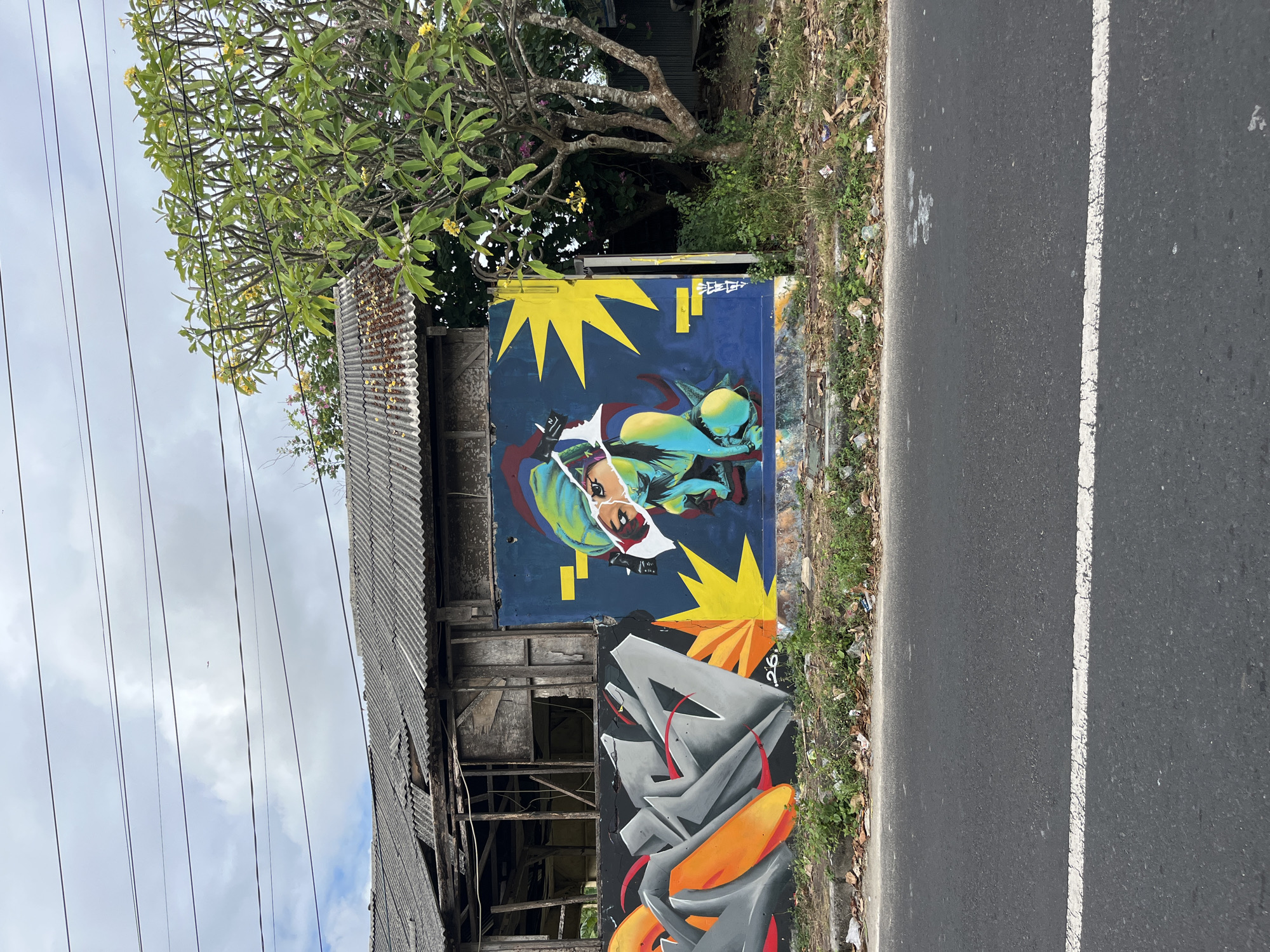 An outdoor scene in Kuta Selatan, Indonesia, features a section of a road with a white line painted in the foreground. Beyond the road, a low wall displays colorful graffiti. On the left, abstract graffiti in shades of gray and orange is visible. To the right of this, a mural on a blue background depicts a person's face rendered in vibrant turquoise, yellow, and red, with a yellow starburst shape behind it. Behind the graffiti wall, an old, dilapidated building with a corrugated roof stands, partially visible. A tree with green leaves and yellow flowers grows to the right of the building, and the sky above is overcast with visible power lines.