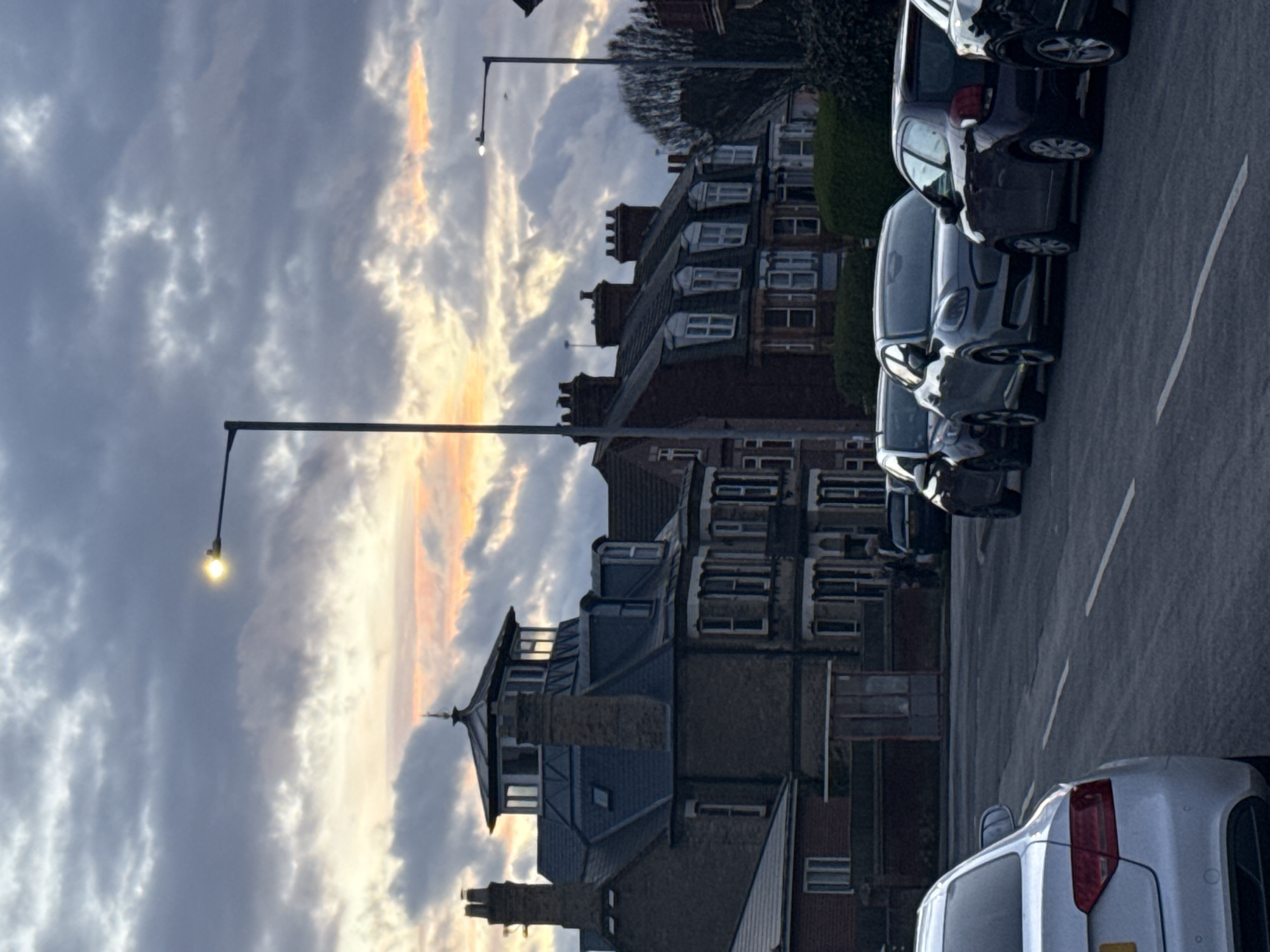 The image shows a street in Gorleston-on-Sea, United Kingdom. Buildings line the street, with parked cars along the curb. The sky is cloudy with a hint of orange light near the horizon, potentially indicating sunrise or sunset. Street lamps are visible. The houses are a mix of brick and stone. A white car is visible in the foreground. The scene is relatively still and calm.