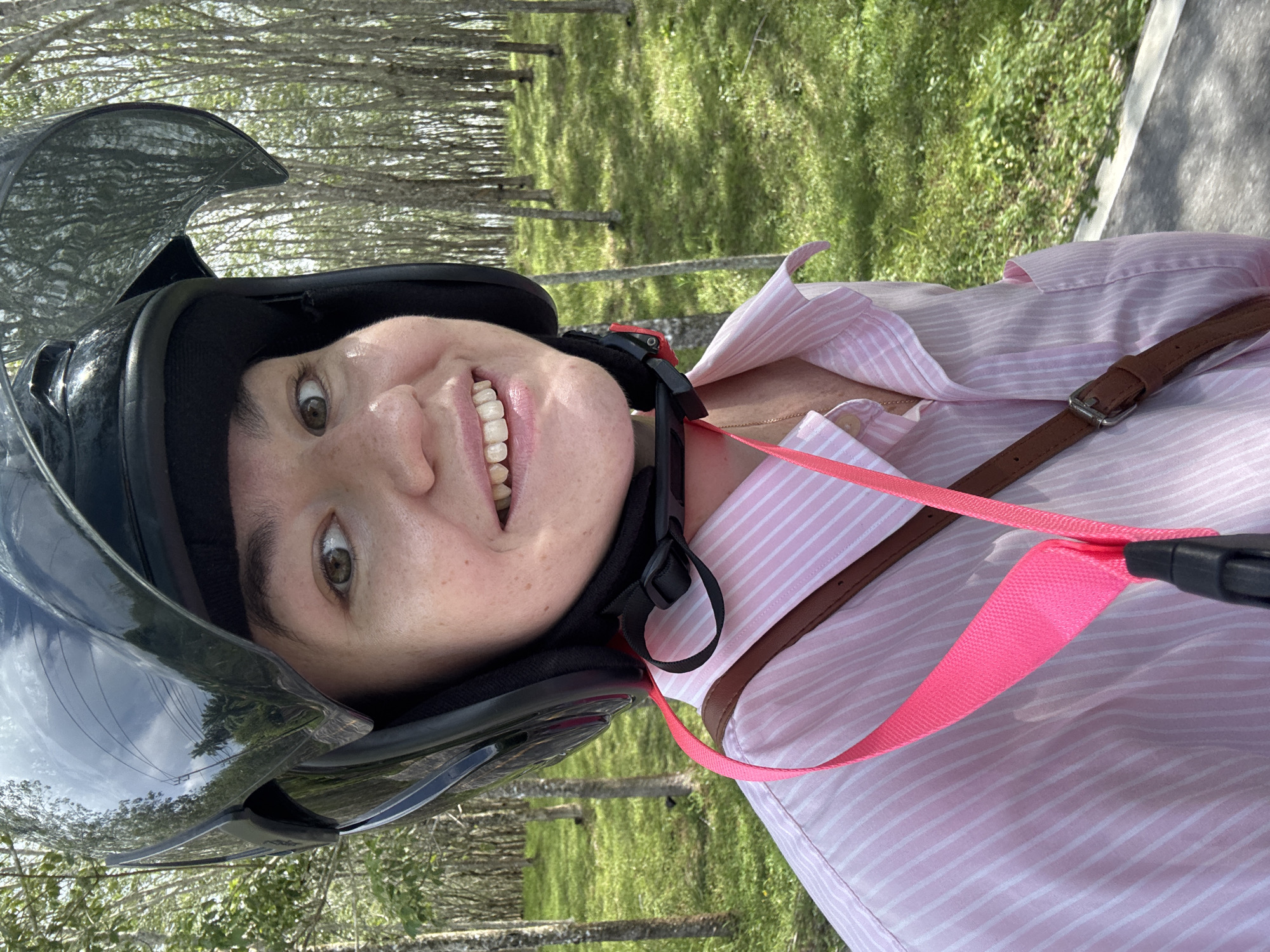 A person wearing a black helmet and a pink and white striped button-up shirt smiles at the camera. A bright pink strap and a brown leather strap are visible across the shirt. The person is outdoors, with a blurred background of trees with thin trunks and green grass. This scene is likely in Pa Khlok, Thailand, a region known for its rubber tree plantations.