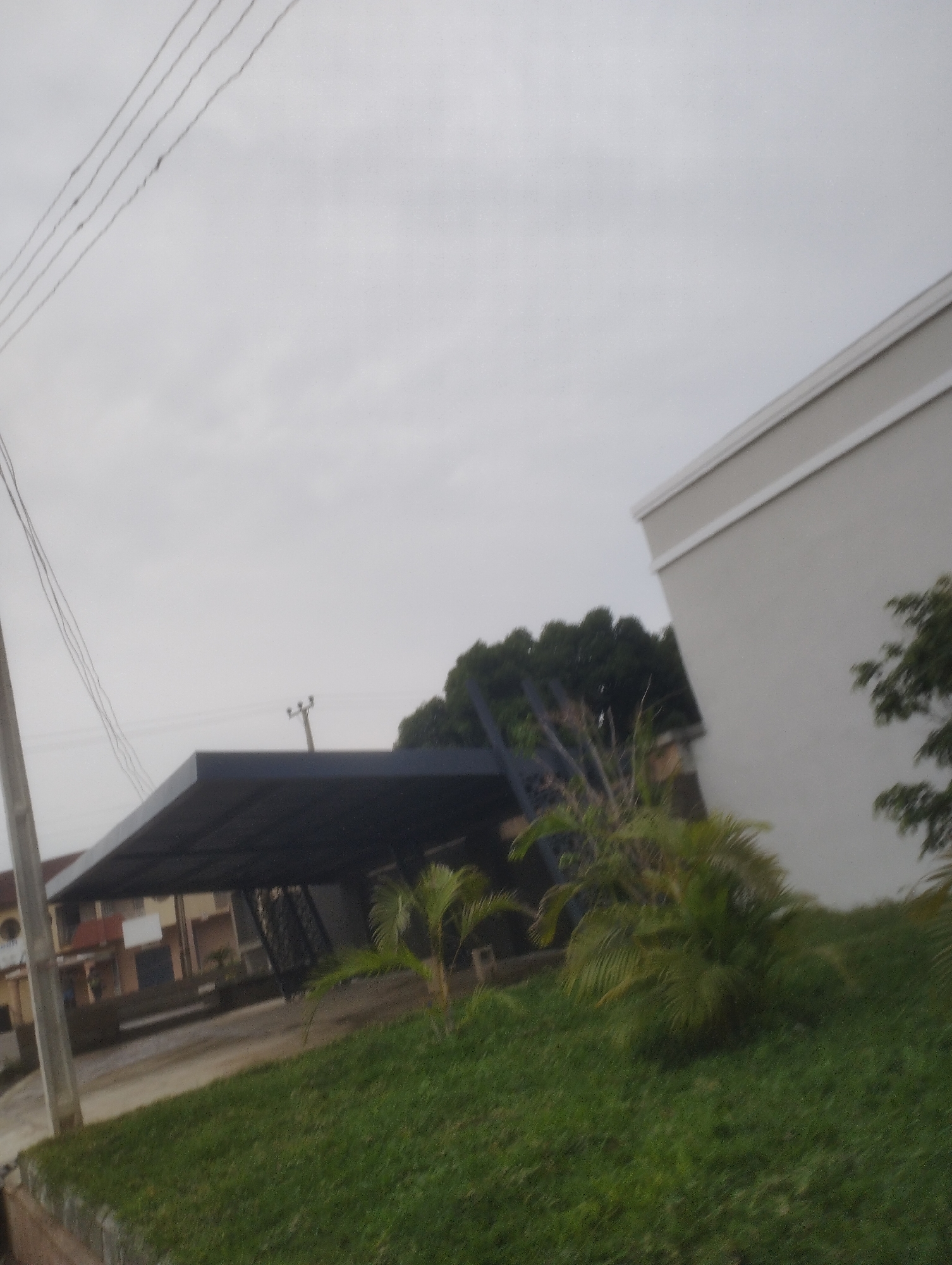 The image depicts an overcast sky over a residential area. In the foreground, a patch of green grass covers a slight incline. Behind the grass, a dark, slanted carport structure is visible. Beyond the carport, a modern, light-colored building with white trim dominates the right side of the frame. To the left of the carport, a multi-story residential building with an orange roof and beige walls can be seen. In the background, dense green trees form a backdrop against the muted sky, and a utility pole with wires is present. The overall scene suggests a quiet, suburban environment, possibly in Jos, Nigeria, given the context.