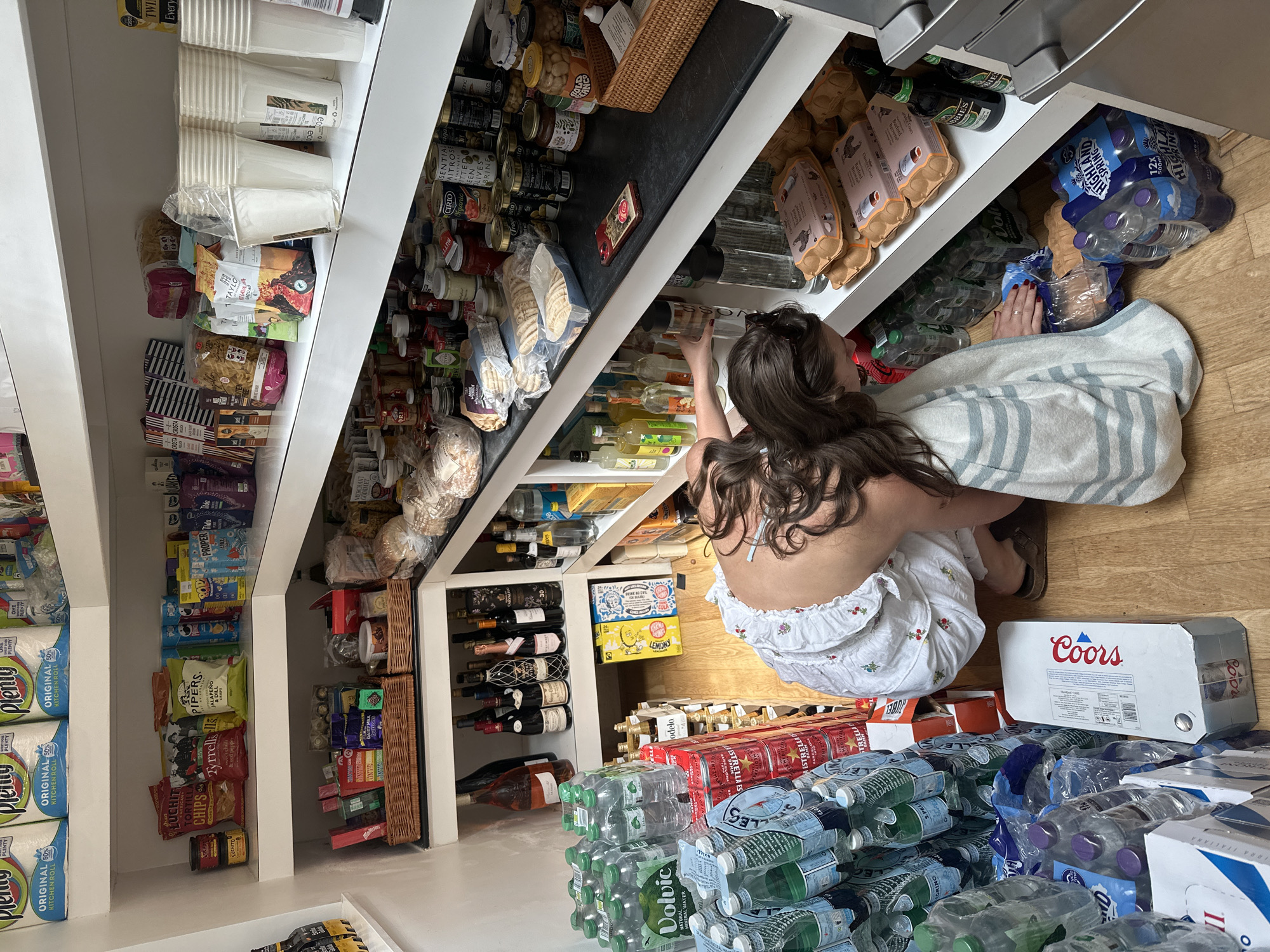 A woman with long curly hair, wearing a white floral dress, is crouched in a walk-in pantry. She is reaching for something on the shelves, which are filled with various food items such as canned goods, pasta, snacks, and wine bottles. The pantry is well-stocked, with multiple cases of bottled water and beer on the floor. Other items visible include paper cups, eggs, kitchen rolls, and various snacks like chips.