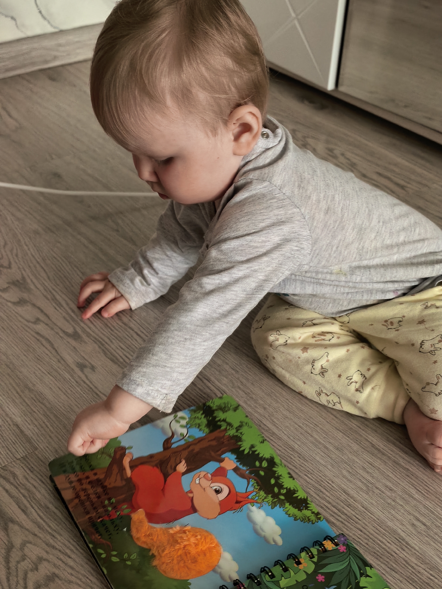 An overhead, slightly angled shot captures a baby or toddler interacting with a colorful book on a light brown wooden floor. The scene is set indoors, likely a home, in Sterlitamak, Russia.
The child, appearing to be about a year old with light-colored hair, is wearing a gray long-sleeved top and yellow pants decorated with numerous white rabbit and star patterns. The baby is positioned on their knees and hands, leaning forward, with their left hand reaching towards the open spiral-bound book. The gesture suggests engagement, possibly turning a page or pointing at an illustration.
The book's front cover is vibrant, featuring a cartoon depiction of a red animal, resembling a squirrel or monkey, swinging from a green tree branch. The background of the illustration shows a bright blue sky with fluffy white clouds. A prominent, soft orange textured patch is affixed to the book cover, adding a tactile element to the design. Faint, unreadable Cyrillic text is visible on the left side of the cover.
In the background, a section of a light-colored cabinet or piece of furniture can be seen. The natural light suggests it is daytime. The overall impression is one of peaceful, focused play or early learning.