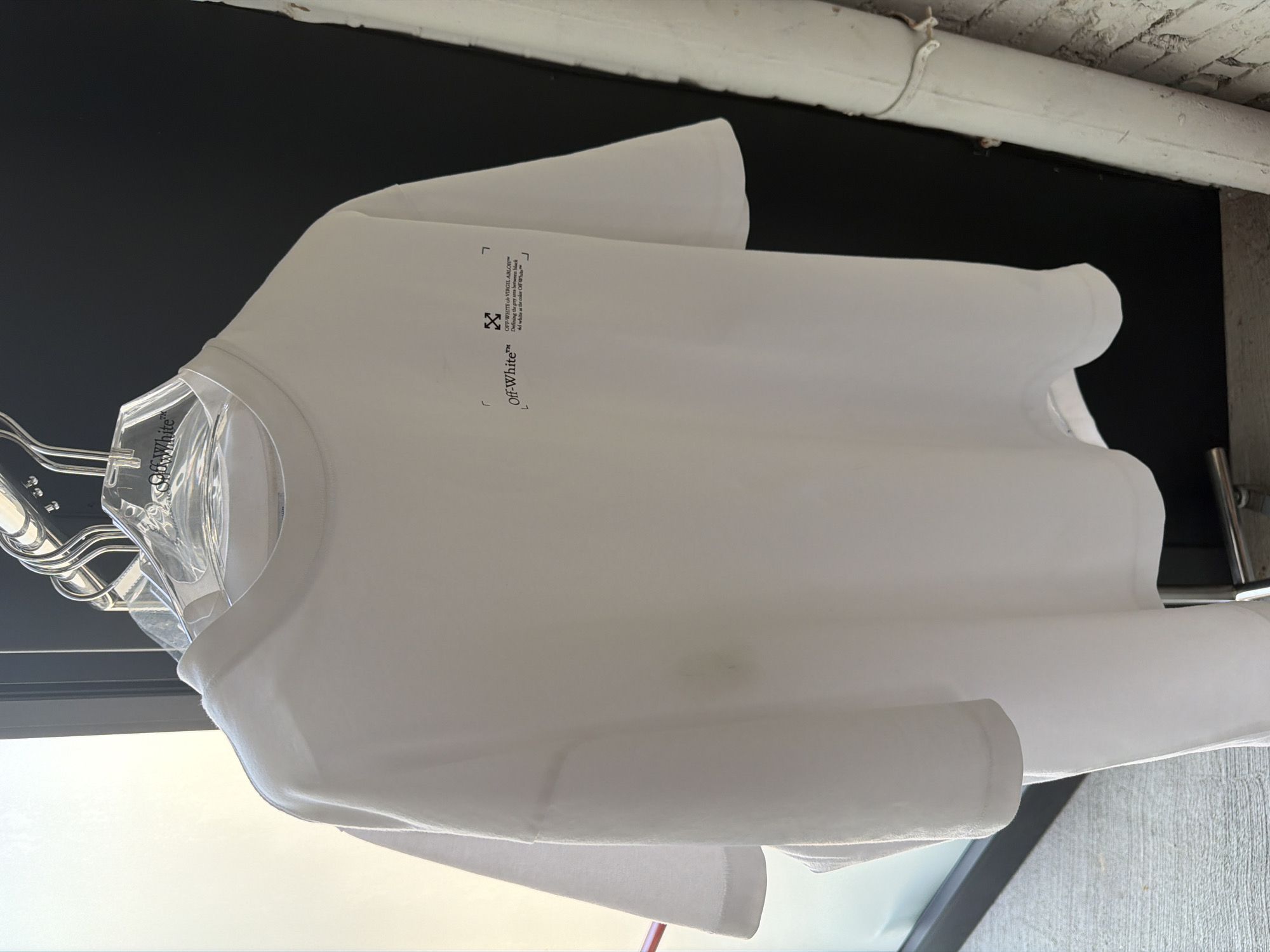A white Off-White brand T-shirt hangs on a clear plastic hanger against a dark background, likely a storefront display. The Off-White logo and brand text are printed on the upper left side of the shirt. The scene appears to be indoors, possibly in a clothing store. It's not possible to determine the exact location in Milan or if it is even in Milan from this image.
