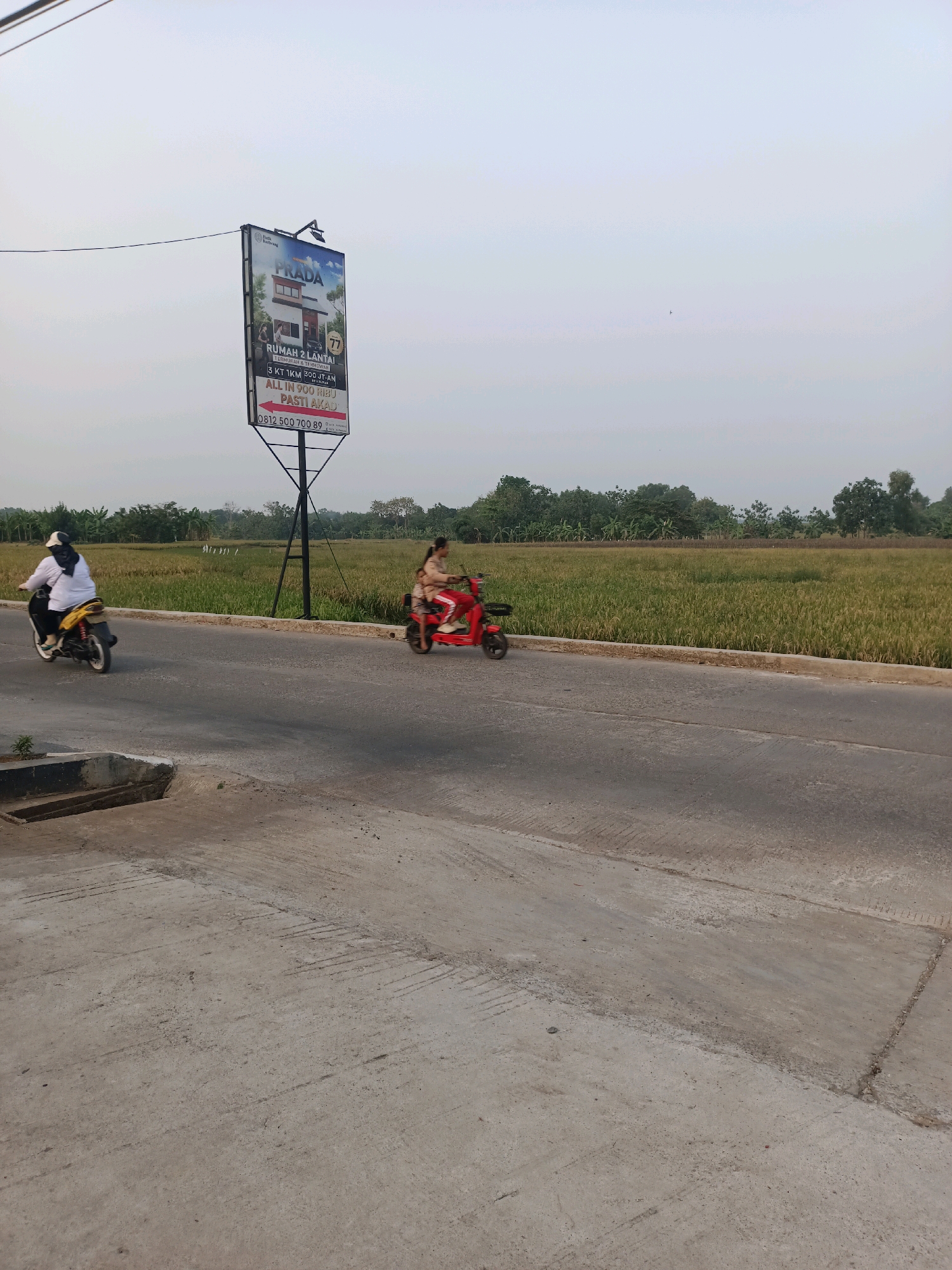 The image captures a roadside scene in Karawang, Indonesia, with a billboard for "PRADA" housing advertisement prominently displayed. The billboard, situated on a metal pole, features an image of a two-story house and text in Indonesian, indicating it is for a "RUMAH 2 LANTAI" (2-story house) that is "TERMURAH & TERBAIK" (cheapest and best). It also mentions "3 KT 1KM 300JT PLAN" (3 bedrooms, 1 bathroom, 300 million Rupiah plan) and an offer of "ALL IN 900 RIBU PASTI AKAD" (all in 900 thousand Rupiah, definitely akad/contract). A phone number, 0812 500 700 89, is also visible.
Two individuals are riding motorcycles on a paved road. The person on the left, on a yellow motorcycle, wears a white shirt and a black and white headscarf. The person on the right, on a red motorcycle, wears a pink top and pants. They appear to be moving away from the viewer.
The environment includes a vast, green field of crops, likely rice paddies, stretching behind the road, bordered by a line of lush green trees. The sky is overcast with a pale, hazy quality. The foreground shows a concrete road with some imperfections and a small drainage channel. The overall impression is one of a semi-rural or developing suburban area on the outskirts of Karawang.