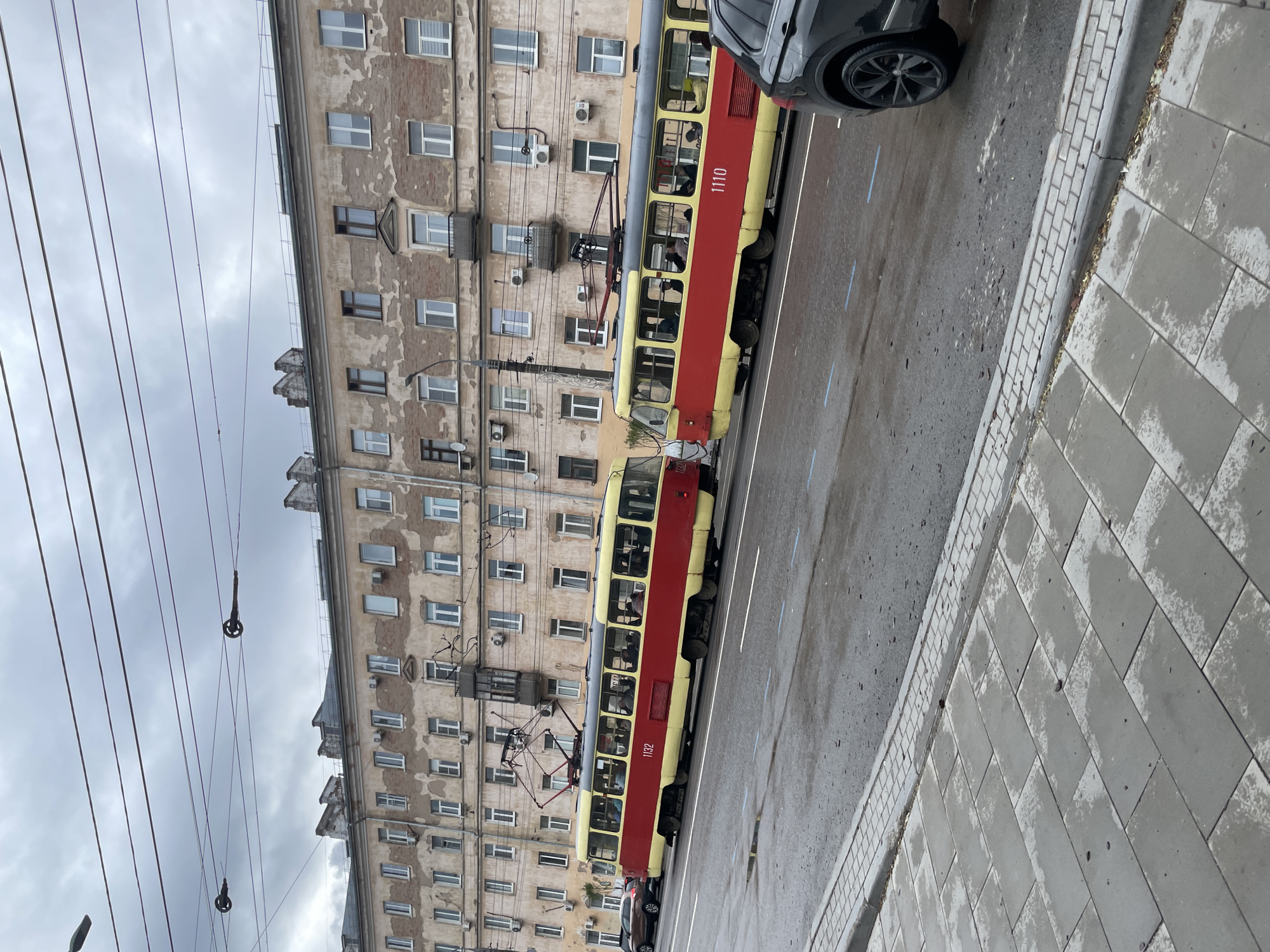 The image depicts a street scene in Izhevsk, Russia. A red and yellow tram, numbered 1110 and 1132, is traveling along the tram tracks in front of a multi-story building with numerous windows and air conditioning units. A black car is partially visible on the street near the tram. The sky is overcast. The sidewalk is paved with gray tiles. Overhead are tram power lines with visible connectors. People are visible inside the tram. The road appears wet, possibly from recent rain.