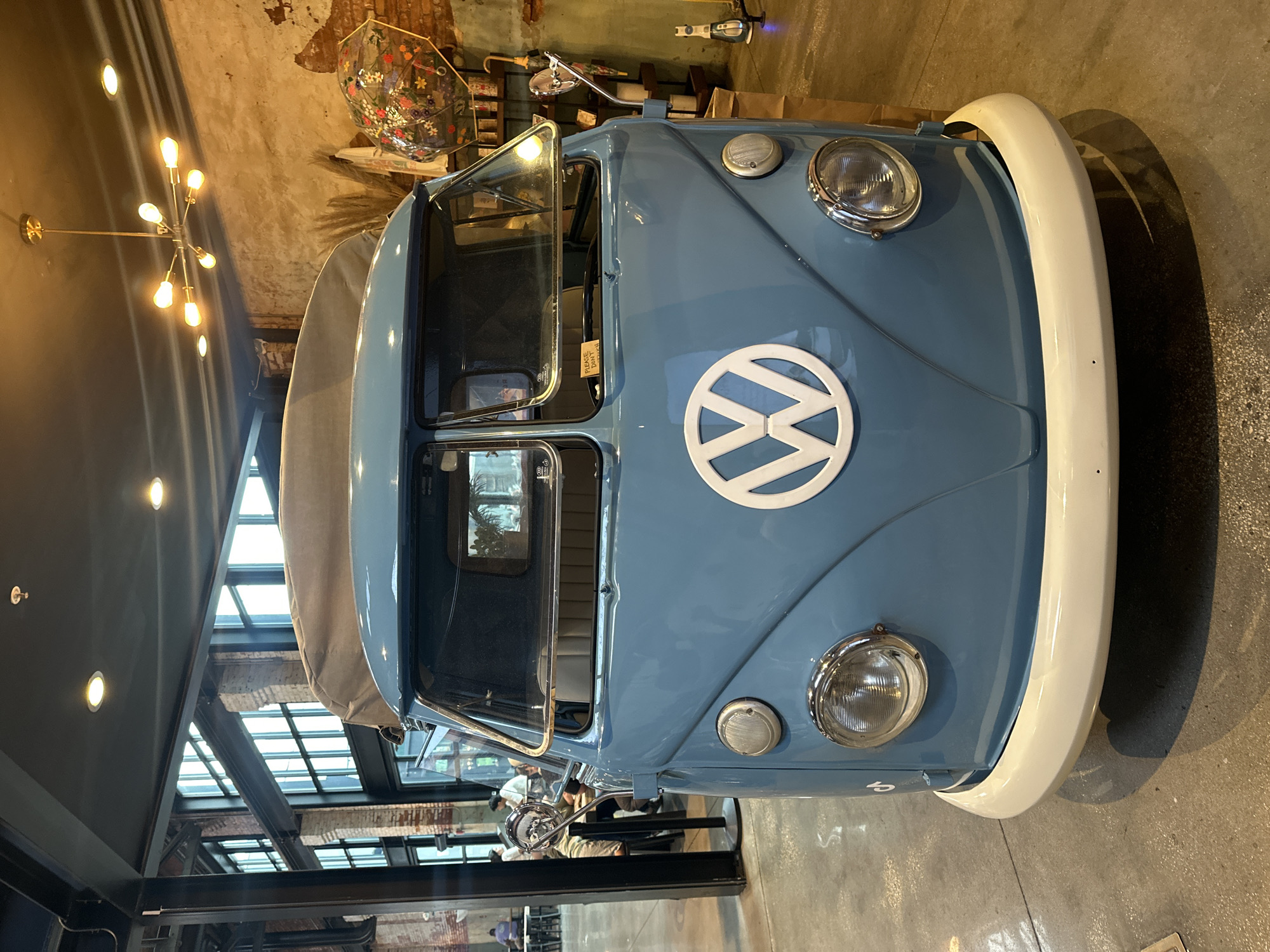 The image shows the front of a light blue Volkswagen van, likely a vintage model, parked indoors. The van features a white VW logo on the hood and a white front bumper. The setting appears to be a restaurant or retail space with large windows and a modern architectural style. A distinctive chandelier is visible in the background, along with other decorative items. Other people can be seen walking near and behind the van. The photo was taken at Heights Public Market at Armature Works.