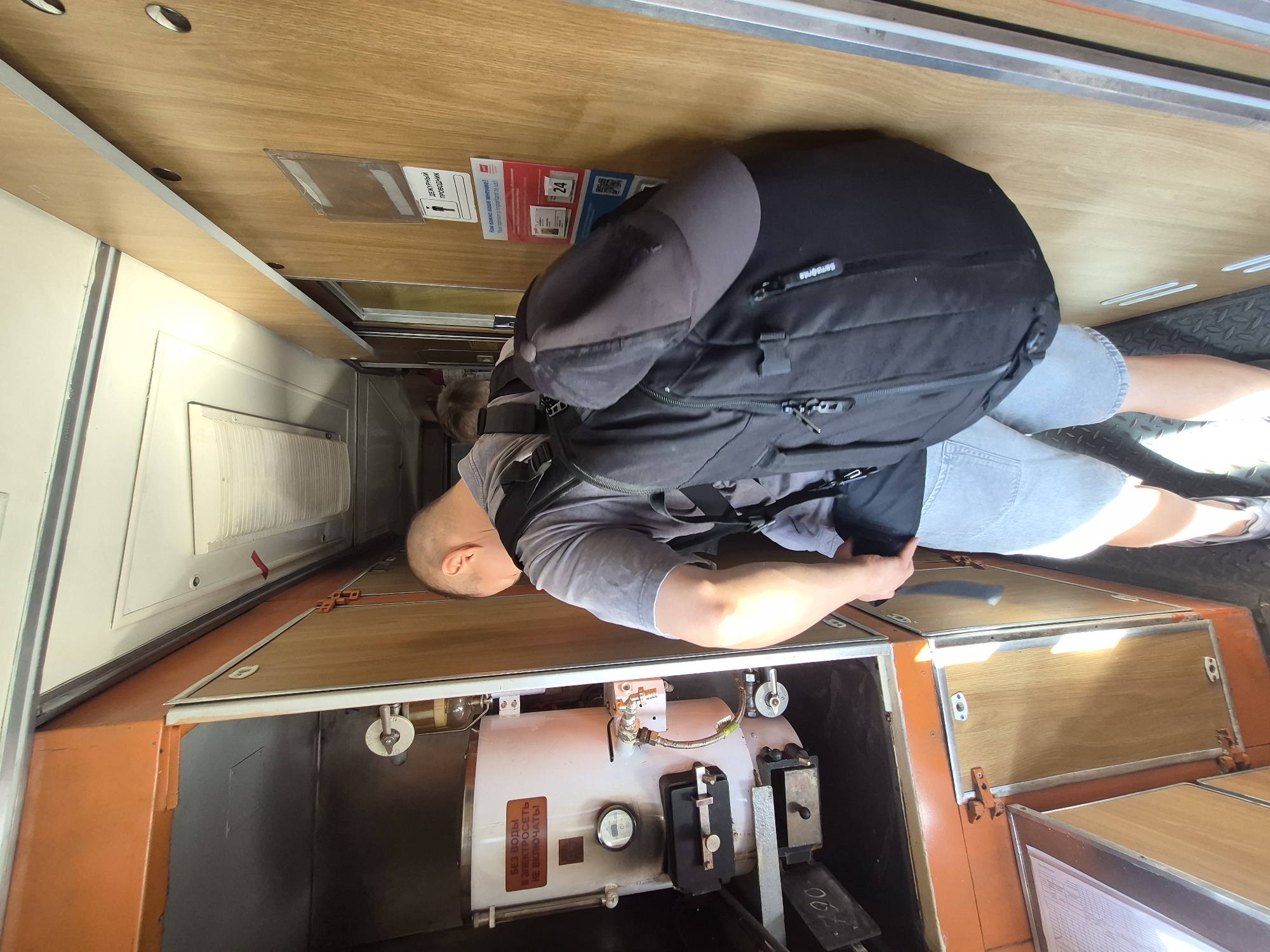 The image depicts the interior of a narrow train car or bus aisle, likely in Yekaterinburg, Russia. A person, viewed from the back, is wearing a grey t-shirt, light-colored shorts, and a large black backpack with a grey baseball cap resting on top. They are standing near a section of cabinetry with an open compartment. Inside this compartment, a white, cylindrical boiler is visible, with a pressure gauge and a warning label in Russian that reads "БЕЗ ВОДЫ В ЭЛЕКТРОСЕТЬ НЕ ВКЛЮЧАТЬ!" (Do not turn on the power without water!). The walls of the compartment and the adjacent corridor are paneled with light-colored wood. There are also some small signs on the wooden wall, one indicating "ДЕЖУРНЫЙ ПРОВОДНИК" (Duty Conductor) with a pictogram of a person. The flooring is a dark, textured material. Another person's leg and foot, wearing white sneakers, are visible in the lower right of the frame, suggesting the viewer is also within the aisle. The lighting appears to be artificial, originating from the ceiling and possibly from an opening in the compartment.