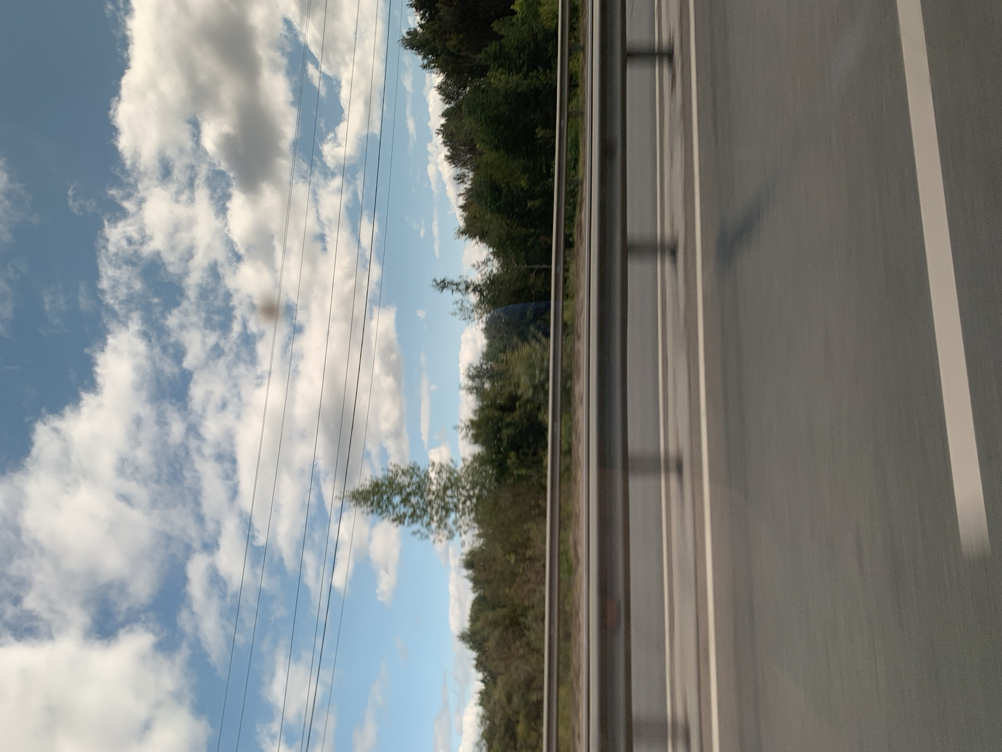 The image shows a view from a vehicle traveling along a highway, with a line of trees bordering the road. The sky is blue with scattered white clouds. There are overhead power lines visible in the upper portion of the image. The highway itself is paved and has white lane markings. A metal guardrail separates the road from the tree line. The location in the city of Iskra, Russia cannot be determined from the visual information provided in the image.