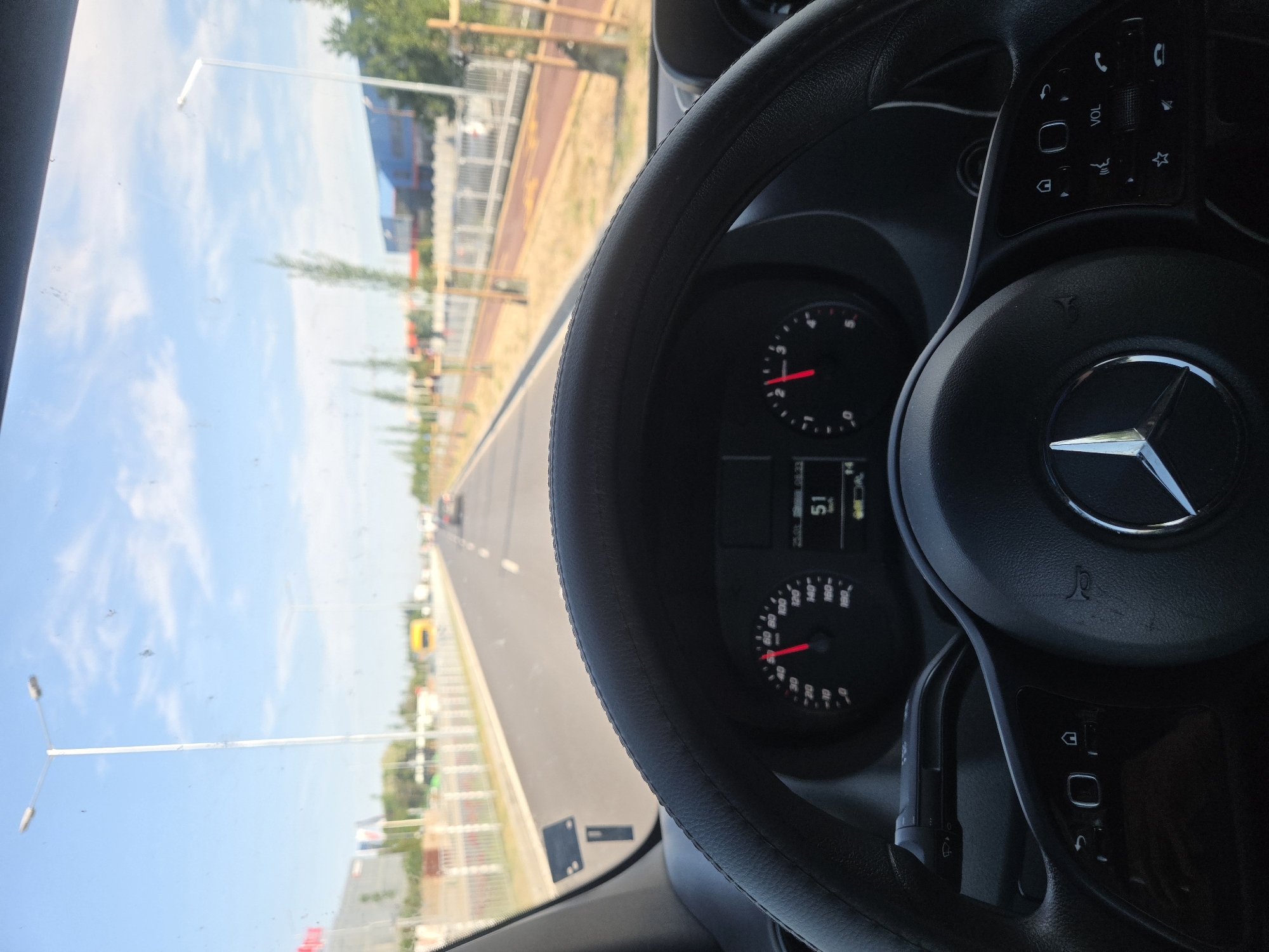 The view is from the driver's seat of a Mercedes-Benz, showing the steering wheel, dashboard instruments indicating a speed of 51, and the road ahead. The road appears to be a paved highway with streetlights and barriers on either side, suggesting an urban or suburban setting. There are trees lining the road, and buildings can be seen in the distance, as well as a clear sky above.
