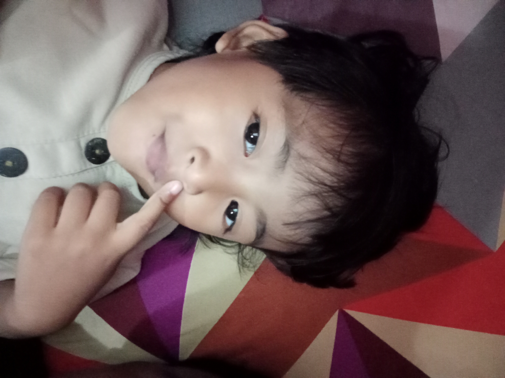 The image shows a close-up, rotated shot of a young child's face and upper body. The child, with dark hair and dark eyes, is lying down and looking towards the camera. Their right hand is raised, with the index finger touching their mouth area. They are wearing a light-colored shirt with two dark buttons visible. The background is a colorful, geometric patterned fabric, possibly a bedsheet or blanket, with sections of red, orange, purple, and beige. The lighting is soft, suggesting an indoor setting. The image's orientation is rotated 90 degrees clockwise. No identifiable Bloro, Indonesia landmarks or context are present in the image itself, therefore its location in Bloro cannot be determined from the visual information provided.