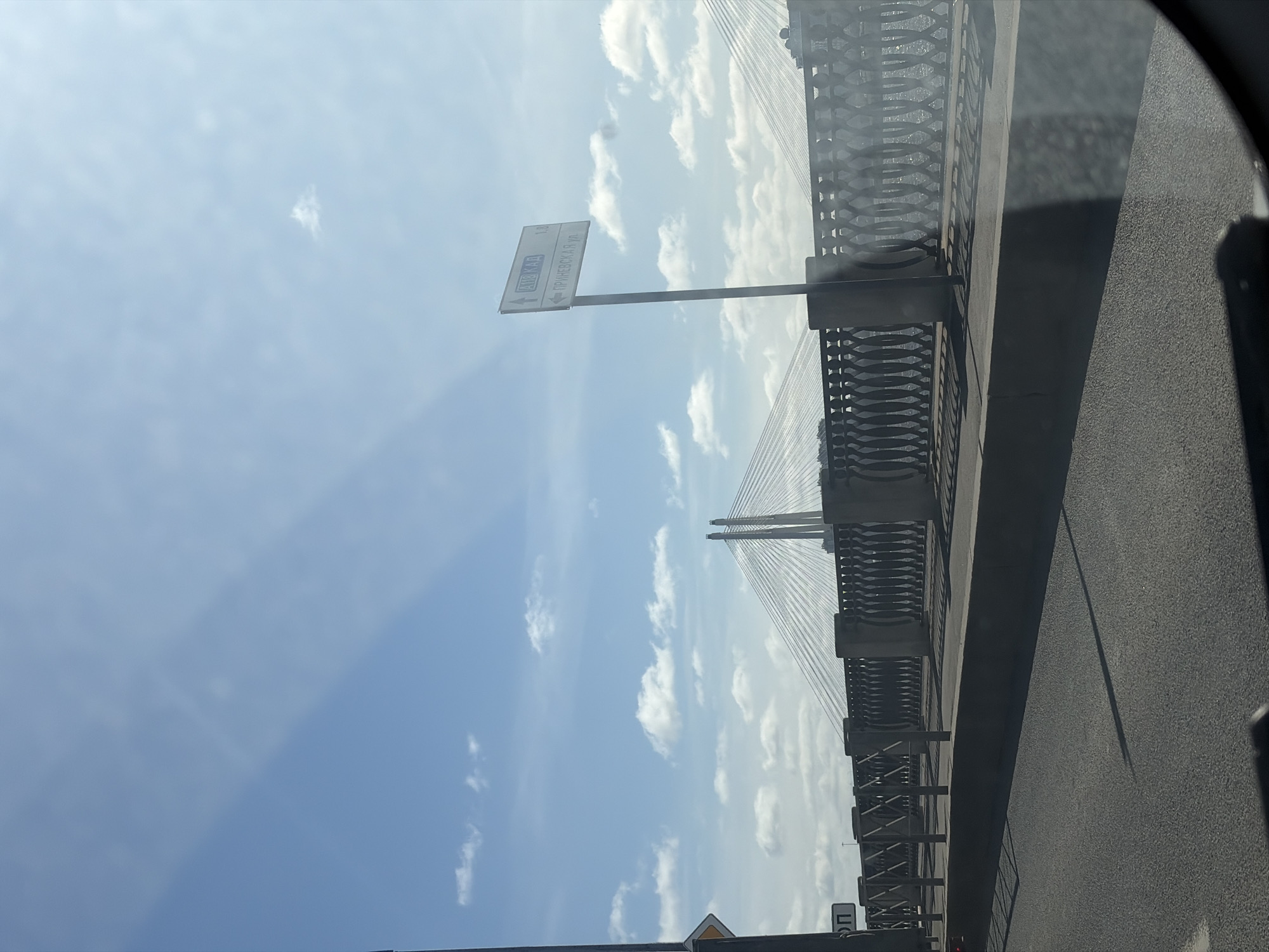 The image depicts a view from a vehicle traveling on the Cable-stayed Bridge in Saint Petersburg, Russia. A street sign indicating directions to "AOBKAA" and "ГР/ИНЕВСК. К.УЛ." is visible. The sky is partly cloudy. The bridge's structure, including cables and railings, is prominently displayed. The road surface is gray.