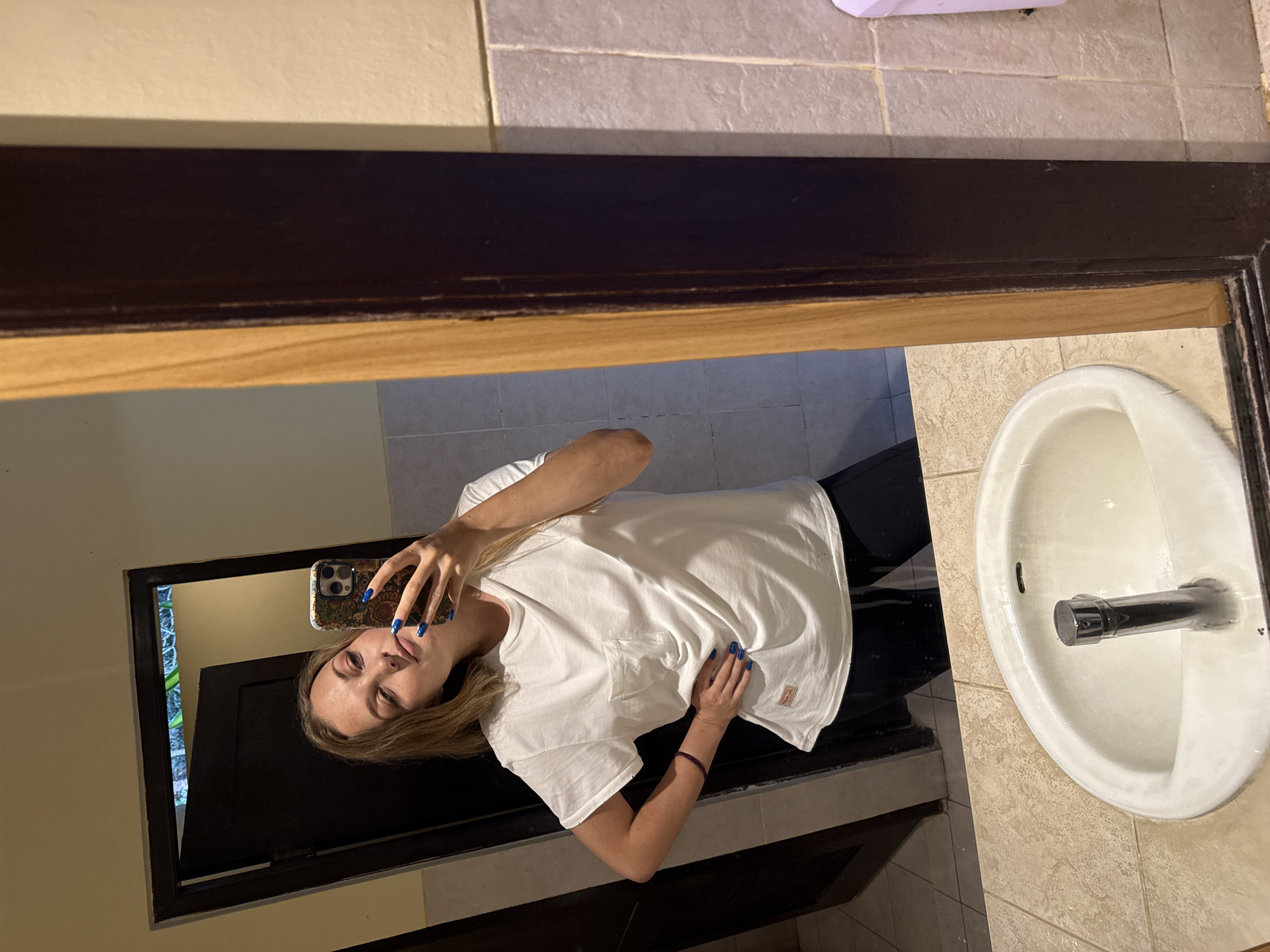 In a dimly lit bathroom, a young woman with blonde hair, wearing a white t-shirt and black pants, takes a mirror selfie. She is standing in front of a large wooden-framed mirror, reflecting her image and the interior of the bathroom. The bathroom features tiled walls in shades of beige and grey, a white sink with a chrome faucet, and a dark wooden door in the background. The woman is posing with one hand on her hip and the other holding her phone, which has a colorful patterned case. Her fingernails are painted a vibrant blue. The scene suggests a casual, personal moment captured in a typical Indonesian bathroom environment, likely in Denpasar, Indonesia.