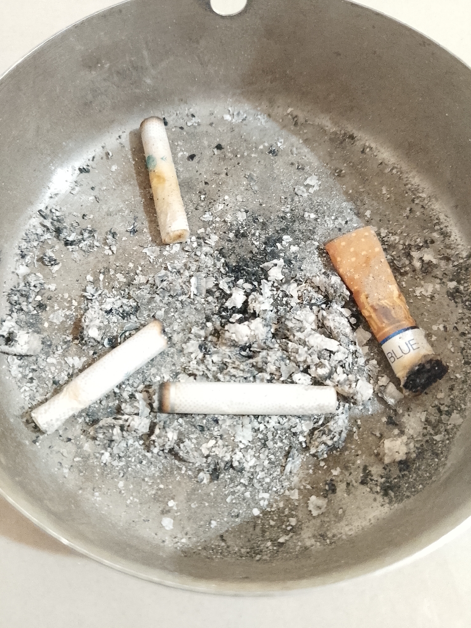 A round, metal ashtray sits on a light-colored surface. The ashtray contains four cigarette butts amidst a scattering of grey ash and debris. One cigarette butt has a brown filter with the word "BLUE" printed on it. The other three have white filters. The image was taken indoors. The location of Lhokseumawe, Indonesia, cannot be verified from this image.