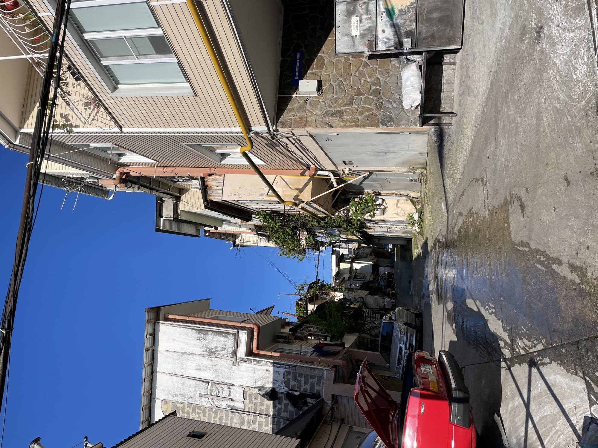 The image captures a narrow, concrete-paved street ascending uphill, identified as Olimpiyskaya Street in Sochi, Russia, by a blue street sign visible on a building on the right. Sections of the pavement are wet, reflecting sunlight.
Multi-story residential buildings line both sides of the street. On the left, buildings display varied construction materials including exposed concrete blocks, white-painted distressed siding, and light-colored horizontal paneling. A brown-orange downspout runs vertically along the facade of one building. In the foreground, the rear of a red sedan is parked with its hood open; its license plate is partially visible, displaying "50occ". Further up the street on the left, a silver sedan with the license plate "у924ун 118" is parked. Laundry is visible hanging on an upper-story balcony on the left side further down the street.
On the right, buildings feature light beige horizontal siding, white-framed windows, and balconies with white railings and potted plants. A prominent yellow gas pipe runs horizontally across the building facade before descending vertically. The ground floor of this building is faced with dark, irregular stone masonry. A gray metal roll-up garage door is adjacent to a darker metal gate, which has partially legible Cyrillic text including "ГАРАЖ" (GARAGE) and "САНЬКА". A simple dark metal bench sits beside the gate, holding a white plastic bag. An electrical box is mounted on the stone wall. Overhead, multiple black utility cables stretch across the street. The sky is clear and intensely blue, indicating bright daylight and casting strong shadows. No people are visible in the scene.