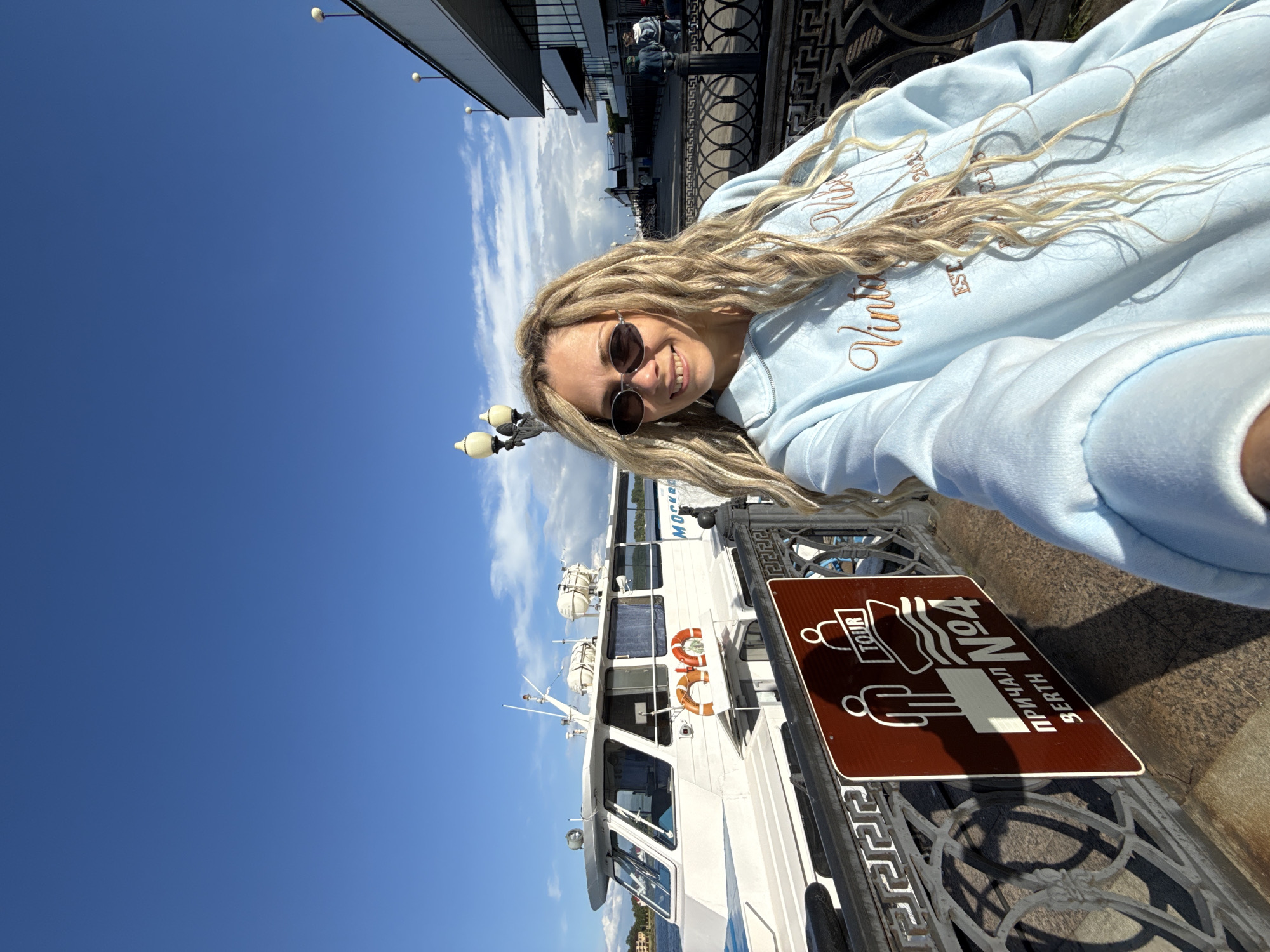 A woman with blonde dreadlocks and sunglasses is taking a selfie in what appears to be Заволжье, Russia. She is wearing a light blue sweatshirt. In the background, there is a white boat with "Moscow" written on it in Russian, along with some orange lifebuoys. Beside her is a brown and white sign with Russian writing and a stylized image of a person on a boat. The sky is blue with some clouds. She is standing by a metal railing.