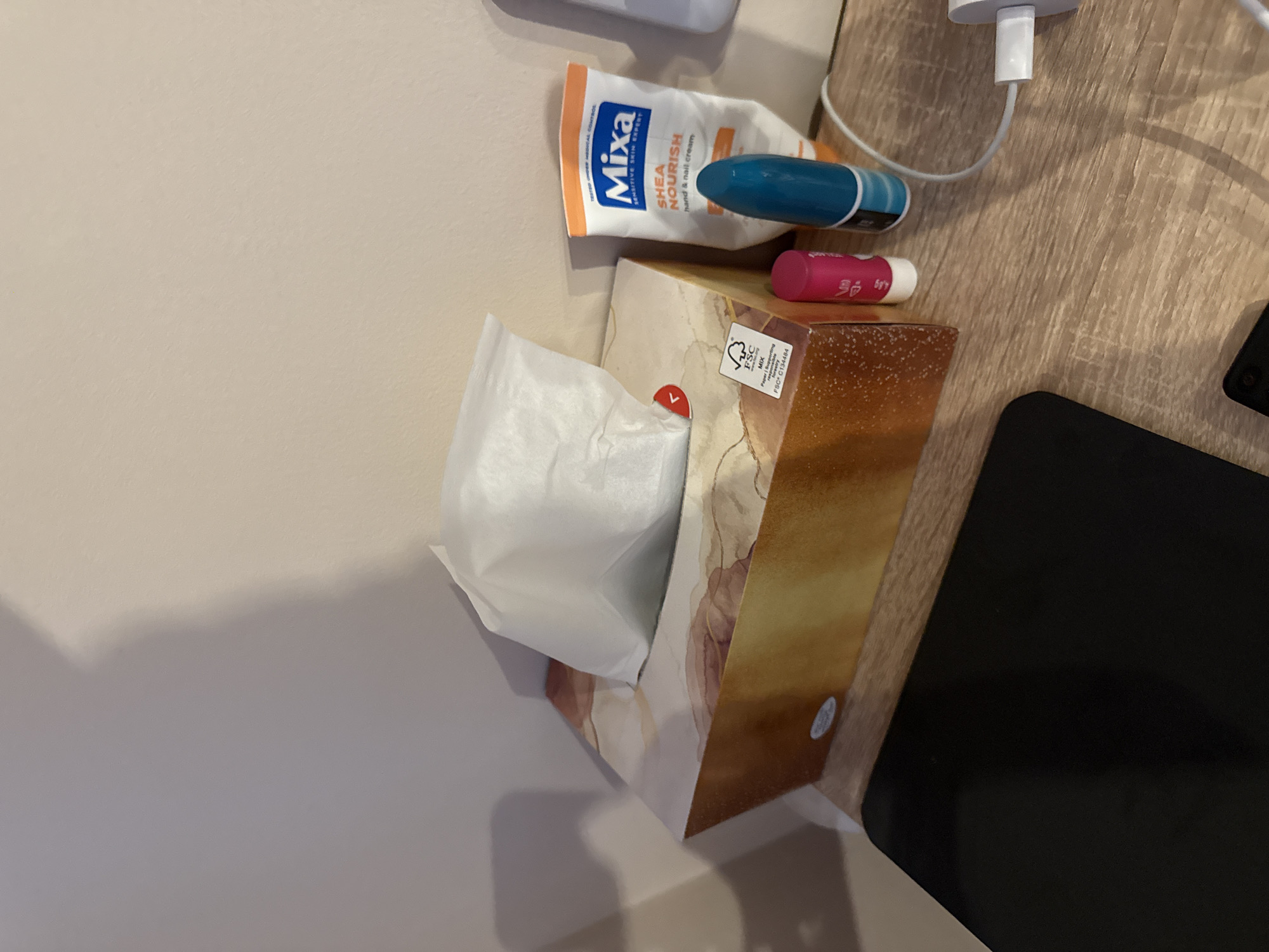 The image shows a top-down view of a bedside table next to a light-colored wall.
On the table, there are several items: a tissue box with a white tissue partially pulled out, a tube of "Mixa Shea Nourish" hand and nail cream, a blue lip balm, a pink lip balm, and a black tablet. Additionally, there's a white charging cable with a block connector. The table surface appears to be a wood-grain laminate.
There is no information in the image or context to indicate a location in the city of Lovech, Bulgaria.