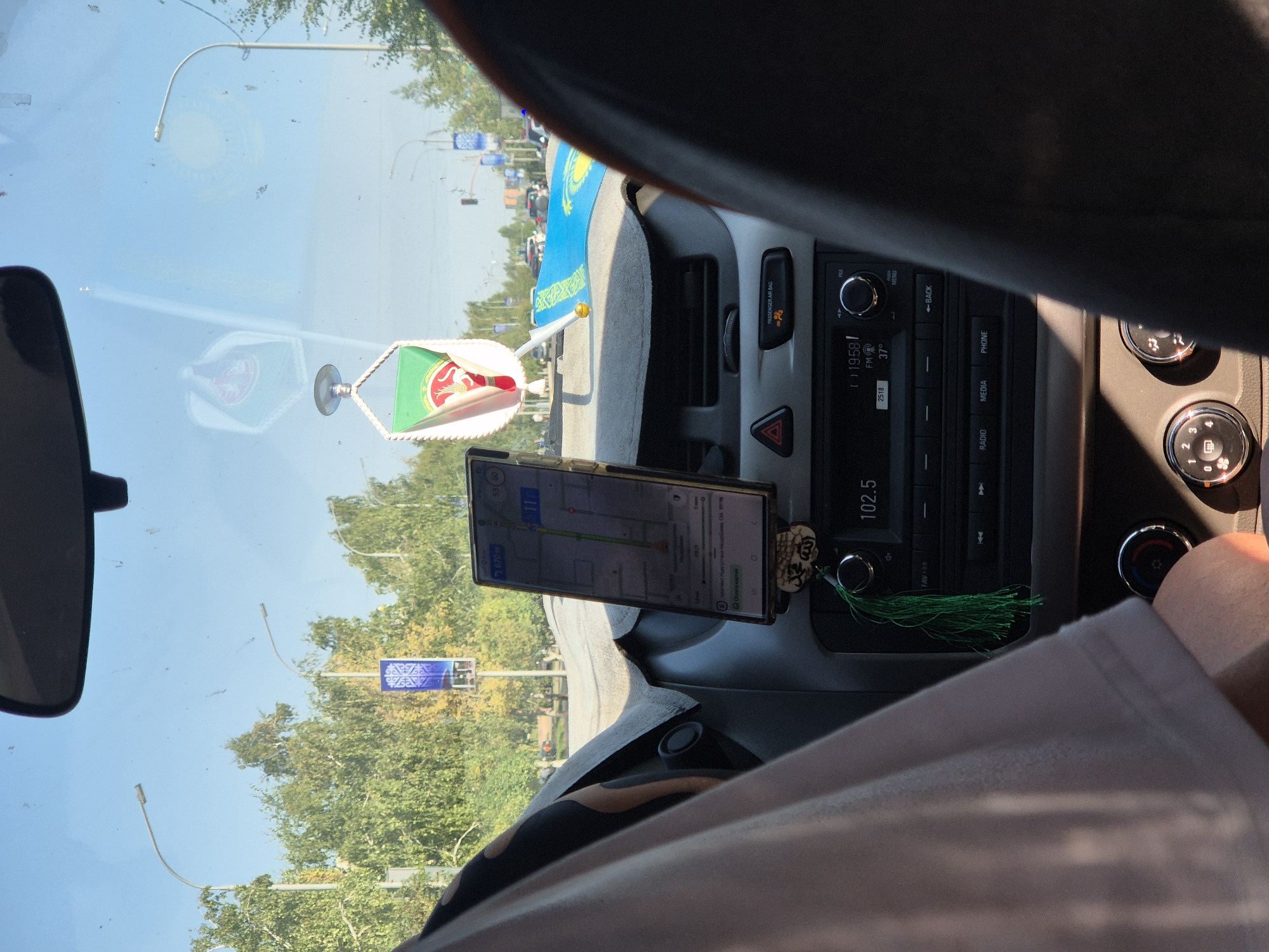 The image shows the interior of a car, viewed from the passenger seat, looking through the windshield. A smartphone is mounted on the dashboard, displaying a navigation app with a route highlighted. Various car controls and a radio are visible. A small flag, possibly of Kazakhstan, is attached to the car's interior. Outside, the scene depicts a city street with trees lining the road and streetlights. The sky is clear and blue. The timestamp on the radio indicates it is 19:58, and the temperature is 37 degrees. The navigation app displays a distance of 670 meters to the destination. The scene is set in Kokshetau, Kazakhstan, as suggested by the presence of the Kazakh flag and the likely context of the image.
