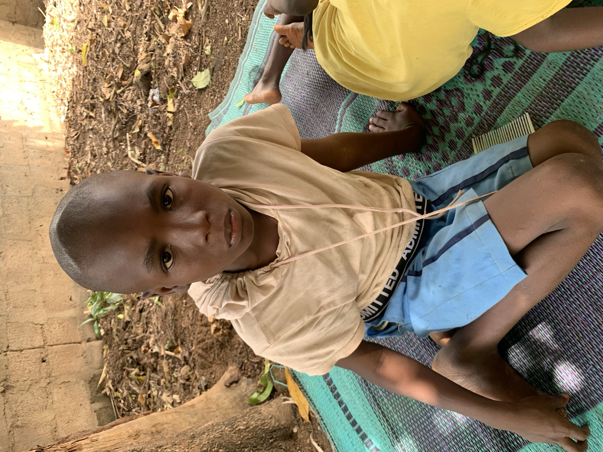A medium, eye-level shot shows a young person in Akwanga, Nigeria, seated cross-legged on a green and purple patterned mat, angled toward the camera. The person has dark skin, a shaved head, and wears a tan short-sleeved shirt and blue shorts with "LIMITED EDITION" printed on the waistband. A thin beige cord hangs around their neck. Their gaze is directed at the camera.
To the right of the person, another individual in a yellow garment sits on the same mat. A comb is placed on the mat near the first person's legs.
To the left, the ground consists of dry leaves and mulch adjacent to a rough, tan brick wall. A portion of a tree trunk lies on the ground near the wall. The environment suggests an outdoor setting, possibly a yard.