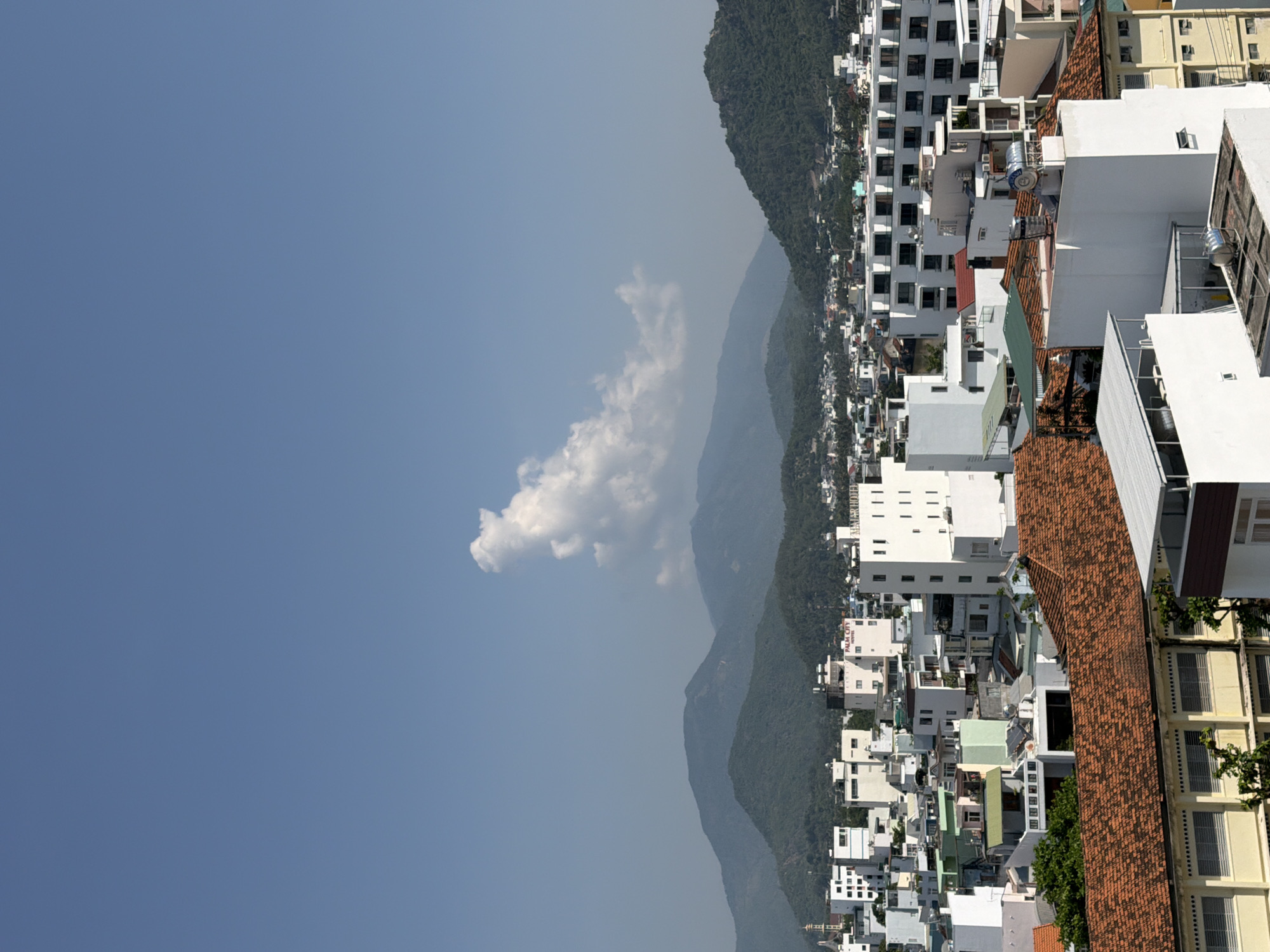 This is a wide shot, taken from a high vantage point, showing a cityscape with mountains in the background. The sky is clear and blue with a single, fluffy white cloud in the upper center of the frame. Below the sky, the image is filled with buildings of various sizes and colors, ranging from residential homes with red-tiled roofs to modern, white multi-story buildings. The buildings are tightly packed together, indicative of a densely populated urban area. In the distance, a range of green, tree-covered mountains forms a backdrop to the city. The overall impression is of a sunny day in a bustling coastal city in Nha Trang, Vietnam.
