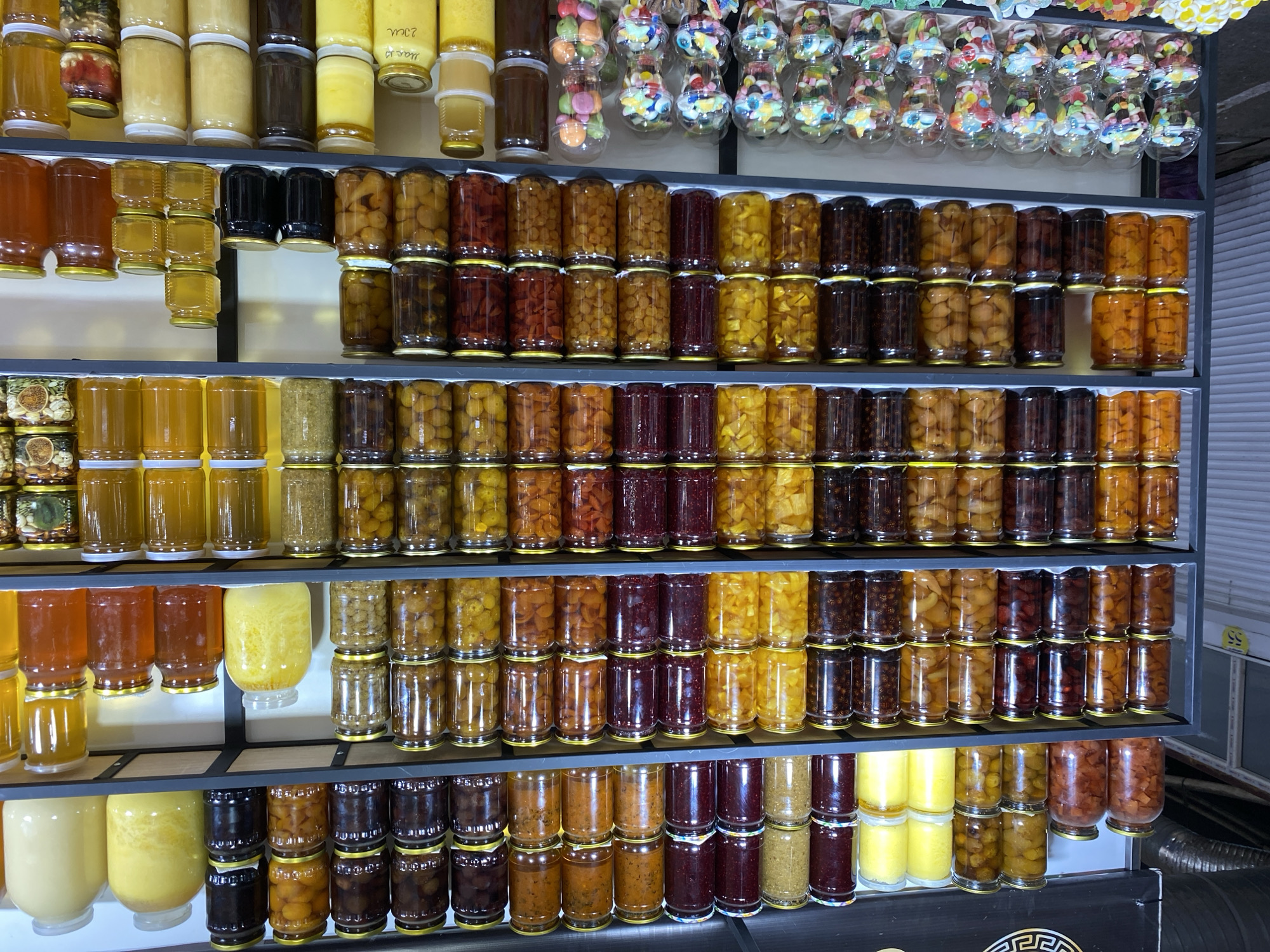 This image displays a street market stall in Makhachkala, Russia, likely within the city's central bazaar or a similar marketplace. The stall is organized with multiple shelves filled with an assortment of preserved goods, primarily fruits and honey, presented in glass jars.
The shelves are made of gray metal, and the background consists of a light-colored textured wall, typical of an indoor market structure. The lighting appears to be a mix of overhead fluorescent lights and ambient light, creating some reflections on the glass jars.
The main subjects are the jars, meticulously arranged in rows. On the top shelf, large glass jars contain light yellow and darker amber colored liquids, possibly honey or preserved fruits. Below, a variety of fruit preserves are visible, including whole fruits, chopped fruits, and jams in shades of red, orange, yellow, and dark brown. The fruit types are not clearly identifiable due to the preservation process.
The middle shelves continue to showcase a similar assortment of fruit preserves. Some jars contain whole fruits like apricots or peaches, while others hold smaller fruits such as berries or cherries. The colors range from vibrant yellows and oranges to deep reds and purples.
The lower shelves feature more fruit preserves and what appear to be jars of honey. Some of these jars are stacked in smaller quantities. There are also clear plastic cups filled with colorful candies and gummy treats, suggesting a section dedicated to sweets.
No people are clearly visible or interacting with the products in the frame. The overall scene is one of a well-stocked and organized food stall, typical of a bustling marketplace. The presence of specific Cyrillic writing on some of the labels, though not clearly decipherable, would further confirm the location as Russia.