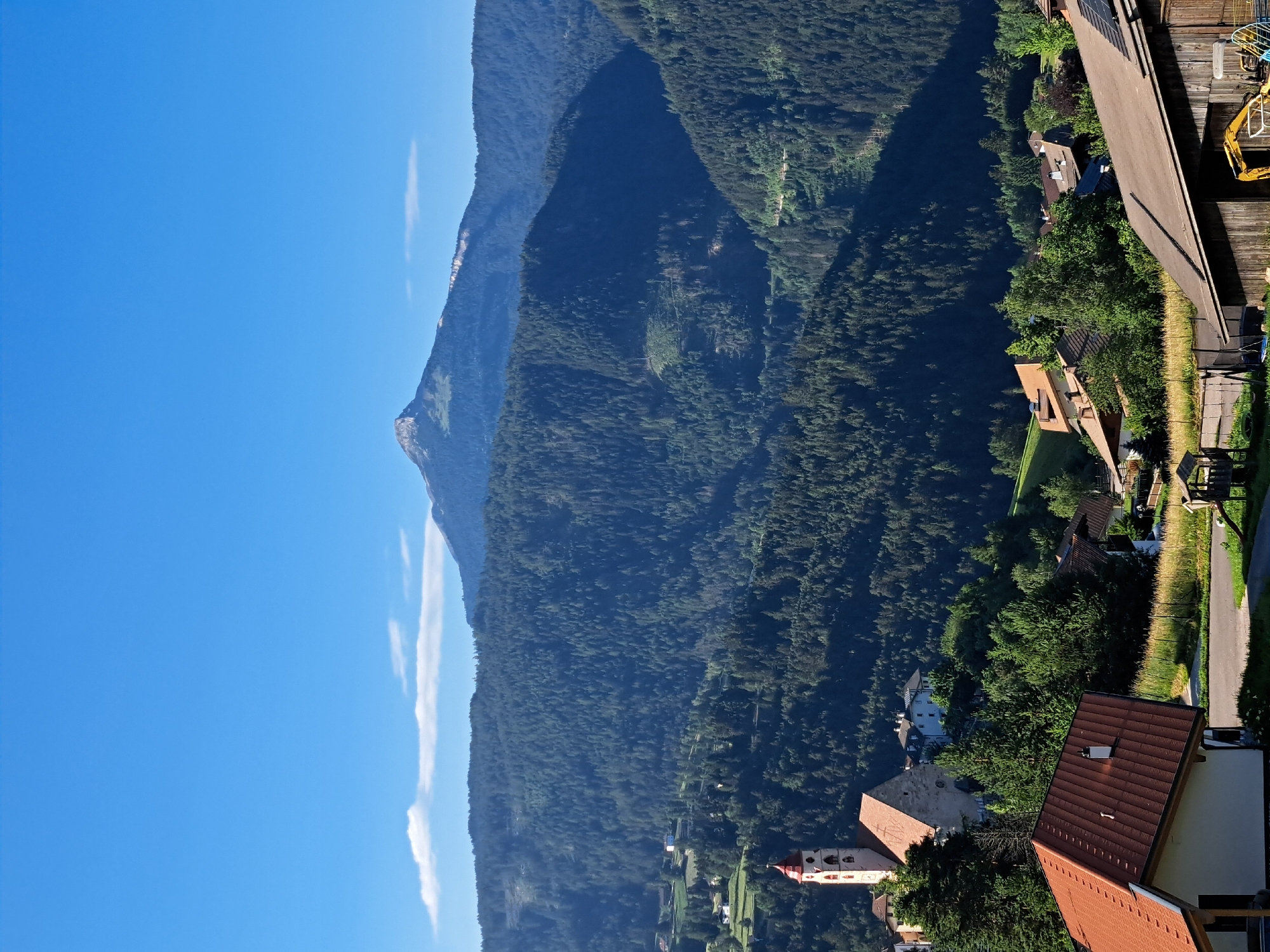 An outdoor, vertical, eye-level, wide-angle photograph captures a sunlit landscape featuring a small village nestled in a valley, with a prominent mountain range in the background. The sky is a clear, bright blue, with a few wispy white clouds.
In the foreground, to the left, a house with a reddish-brown tiled roof is partially visible. To the right of this house, a dirt road is present, with a children's play structure and a trampoline on the grass beside it. Further to the right, a rustic wooden barn-like structure is seen, with a yellow excavator partially visible in front of it.
Mid-ground details include a cluster of houses and buildings, characteristic of a village. A notable structure is a church with a tall, slender bell tower topped with a red roof. The church has a large, reddish-brown tiled roof as well. The village is surrounded by lush green trees and rolling hills.
The background is dominated by a vast, densely forested mountain range. The trees are a deep green, creating a textured, layered appearance. In the far distance, a distinct, pointed mountain peak rises above the forested hills, its slopes partially illuminated by the sun. The overall impression is one of a serene, rural alpine setting.